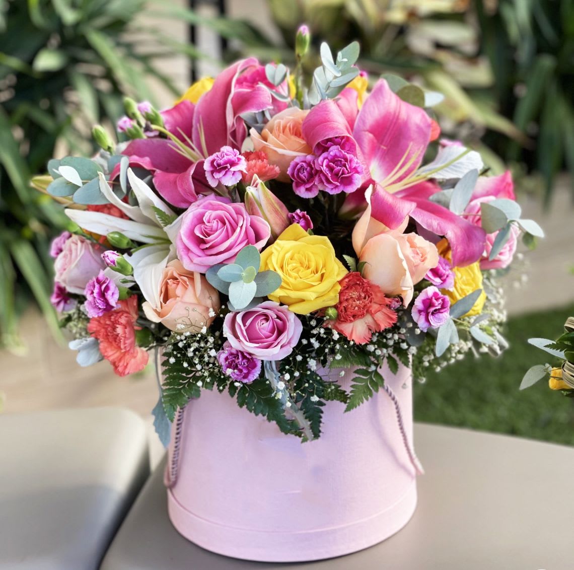 Blushing Garden Bloom Box - A cheerful and radiant expression of love, this garden-inspired arrangement bursts with color and charm. Featuring a delightful mix of pink lilies, yellow and peach roses, lavender and hot pink spray carnations, and lush greenery—this bouquet is nestled in a pastel pink hatbox, perfect for birthdays, celebrations, or simply brightening someone’s day.