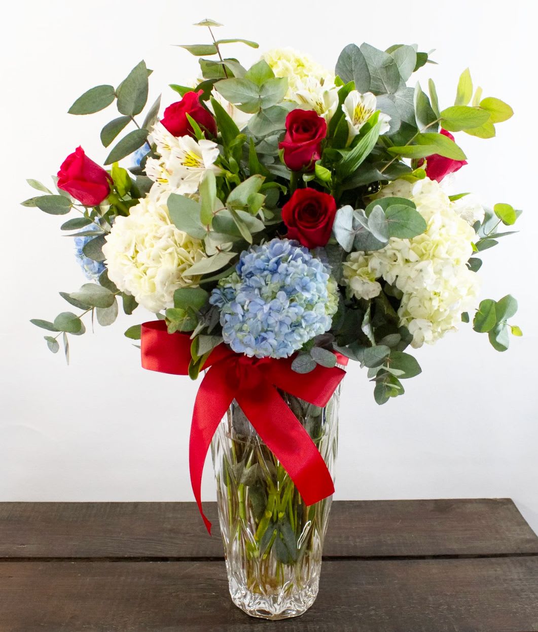 Hydrangea Charm Vase Arrangement - This timeless bouquet features a lush mix of red roses, white and blue hydrangeas, alstroemeria, and eucalyptus greens. Beautifully arranged in a tall, textured glass vase and tied with a striking red satin ribbon, it’s a graceful gift for any occasion—birthdays, thank yous, or simply to brighten someone’s day.