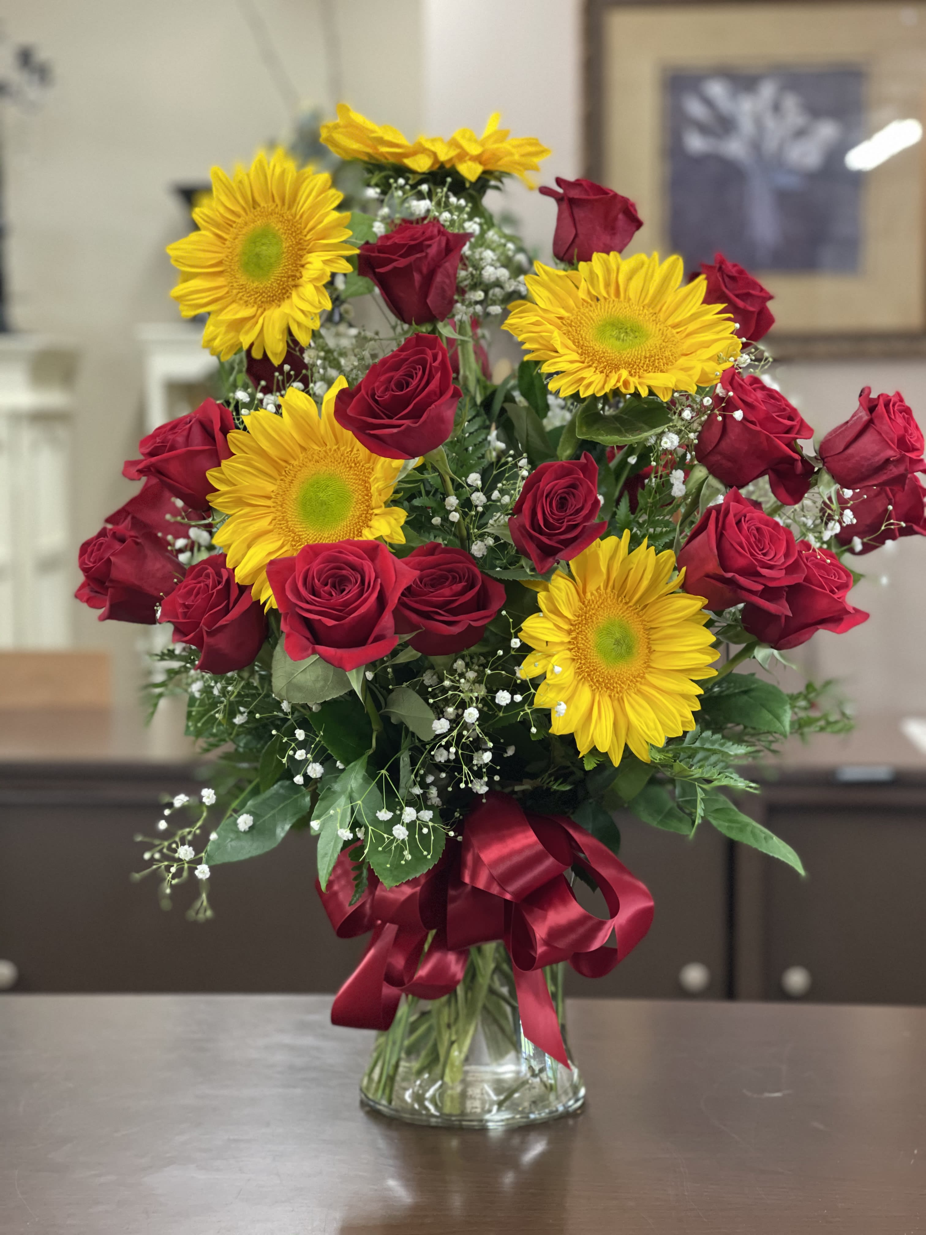 2 Dozen Red Roses with Sunflowers - Bold red roses meet sunny sunflowers in a striking summer-ready bouquet that reads as both romantic and joyful - perfect as an anniversary bouquet, birthday surprise, or a just-because celebration for the person you love. The contrast of deep crimson and golden yellow creates an immediate visual impact; small white filler blooms weave through the arrangement to soften the composition and add light, airy texture that keeps the look fresh for any occasion, including Mother's Day or Valentine's Day.  This flower arrangement includes two dozen red roses arranged with large yellow sunflowers and delicate white filler blossoms, all placed in a clear glass vase and tied with a wide red satin ribbon for a polished finish. The mix of full, velvety rose heads and bold sunflower faces gives a lush, full silhouette that reads as both elegant and exuberant - an ideal gift when you want the recipient to feel adored and celebrated.  Send this as a romantic gesture, a cheerful birthday bouquet, or a bright pick-me-up to congratulate someone special; it's also a standout summer centerpiece. Care is simple: fresh water in the vase and a cool spot away from direct heat will help the blooms last longer so she'll enjoy your thoughtful surprise for days.