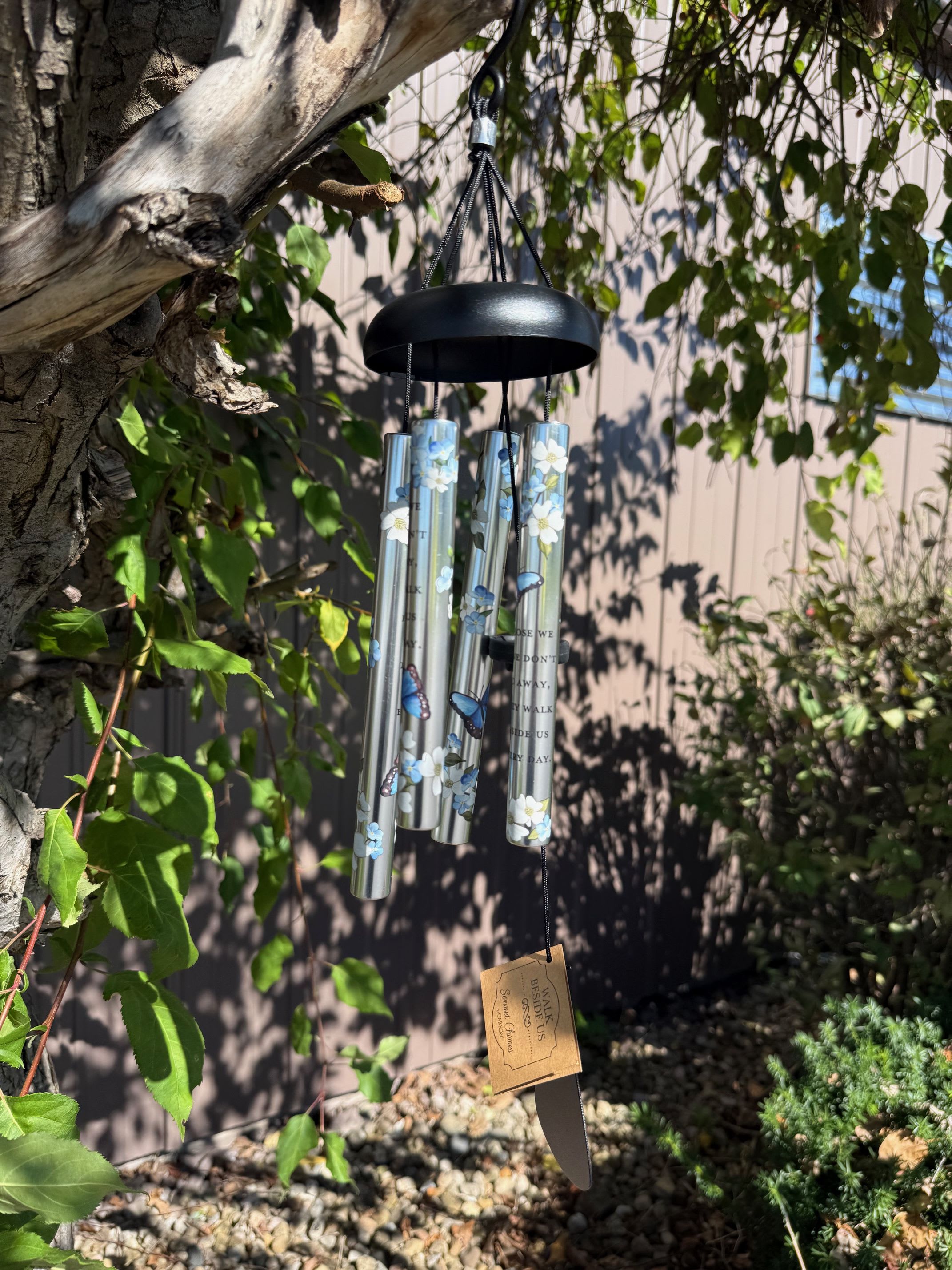 Decorative silver wind chime with blue butterflies and white flowers hanging from a tree branch