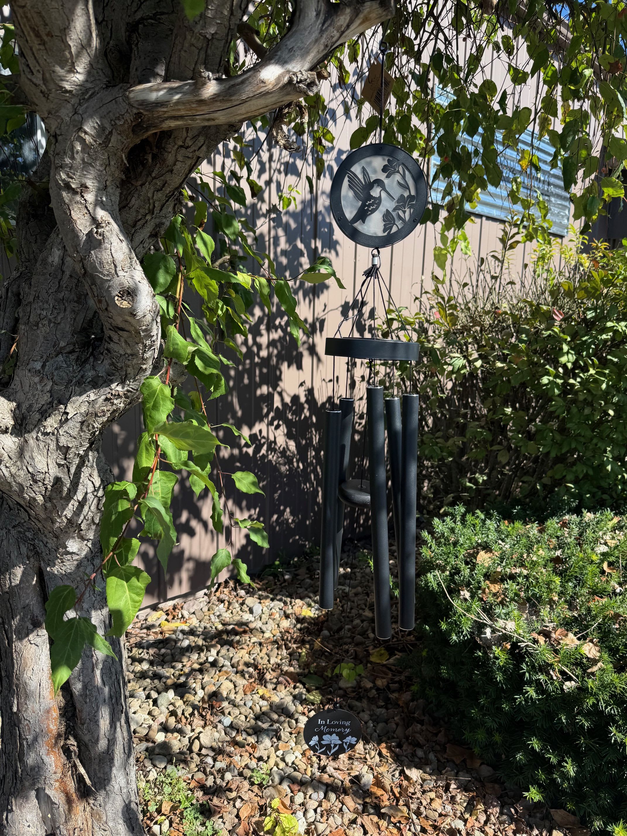 Black memorial wind chime with hummingbird design hanging from a tree over a rock garden.