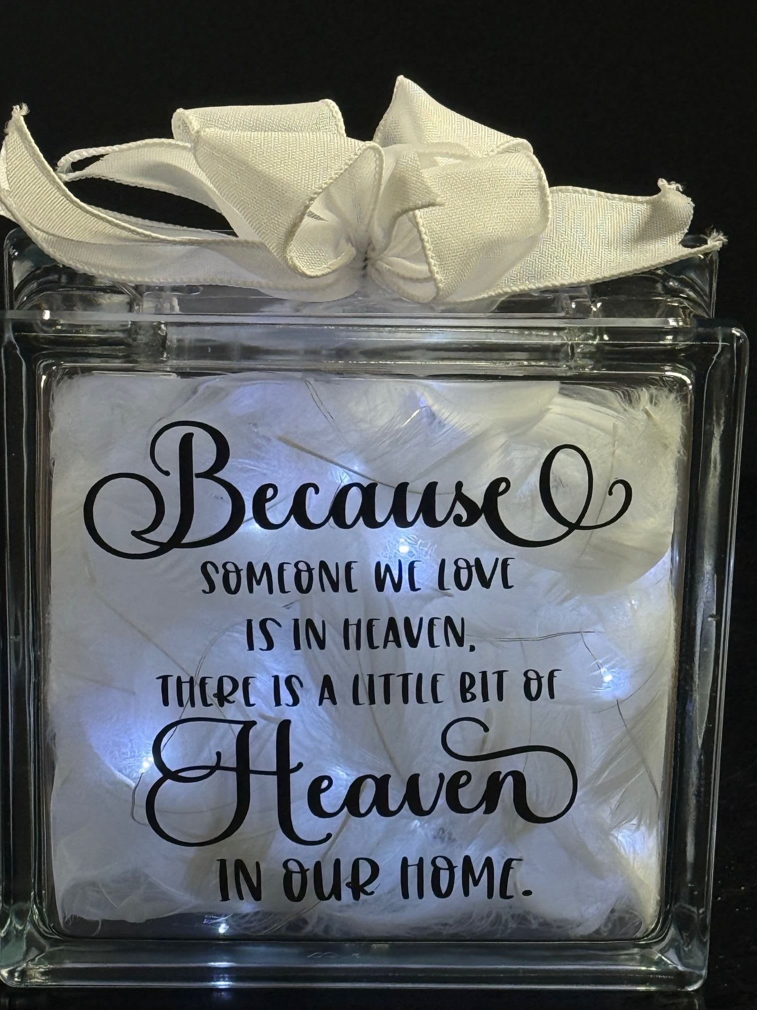 Lighted glass block with white feathers, ribbon, and a memorial quote about a loved one in heaven.