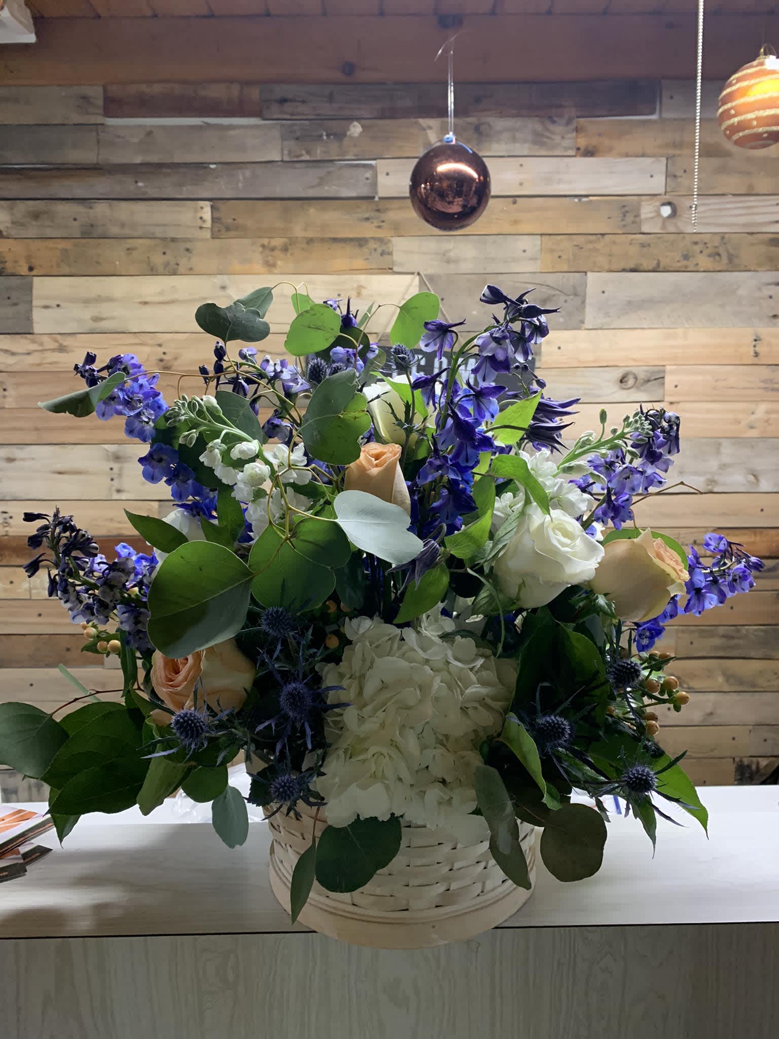 Treasured Basket - The blooms of this beautiful basket filled with  blue, white and peach flowers to brighten anyones day! Flowers may include delphinium, roses, hydrangea and so much more.