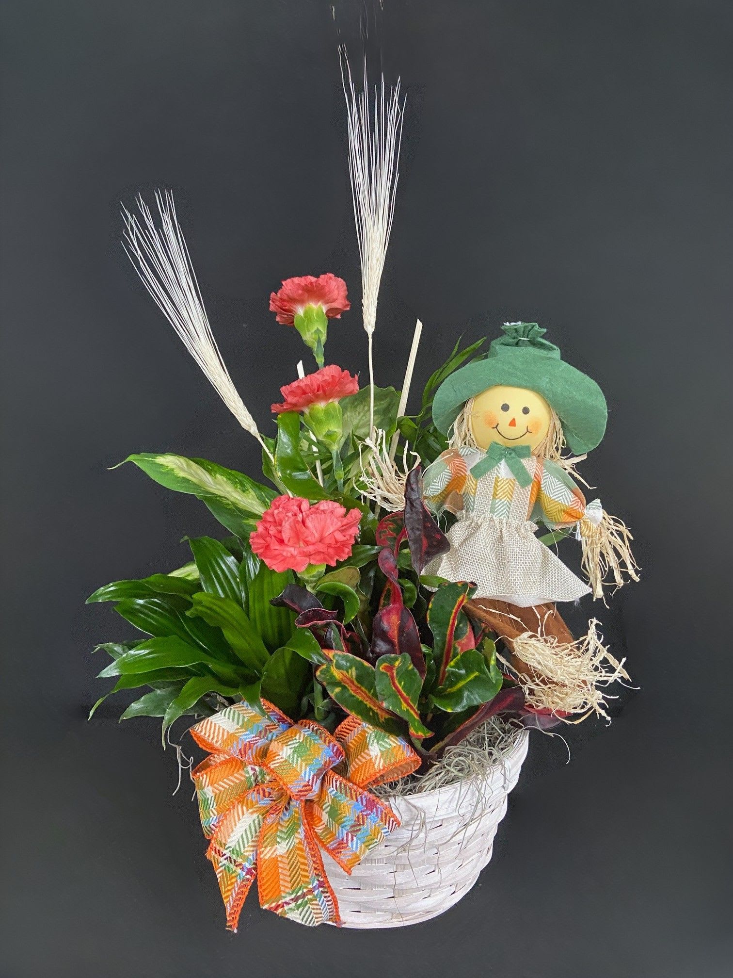 Basket arrangement with pink carnations, tropical foliage, and a scarecrow doll