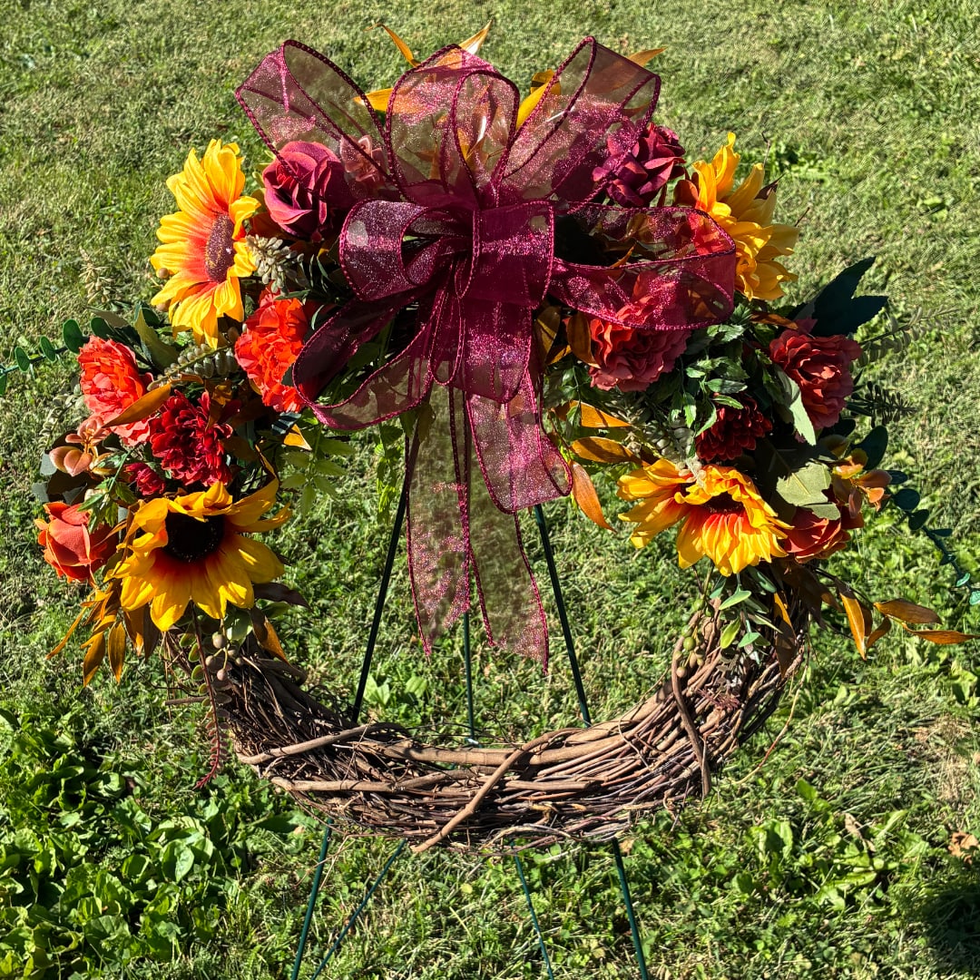 Floral wreath with sunflowers, red blooms, and a burgundy bow on a stand