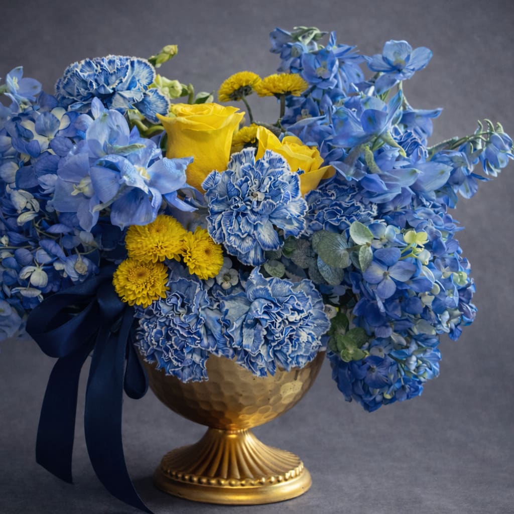 Blue and yellow floral arrangement in a gold vase with a navy ribbon
