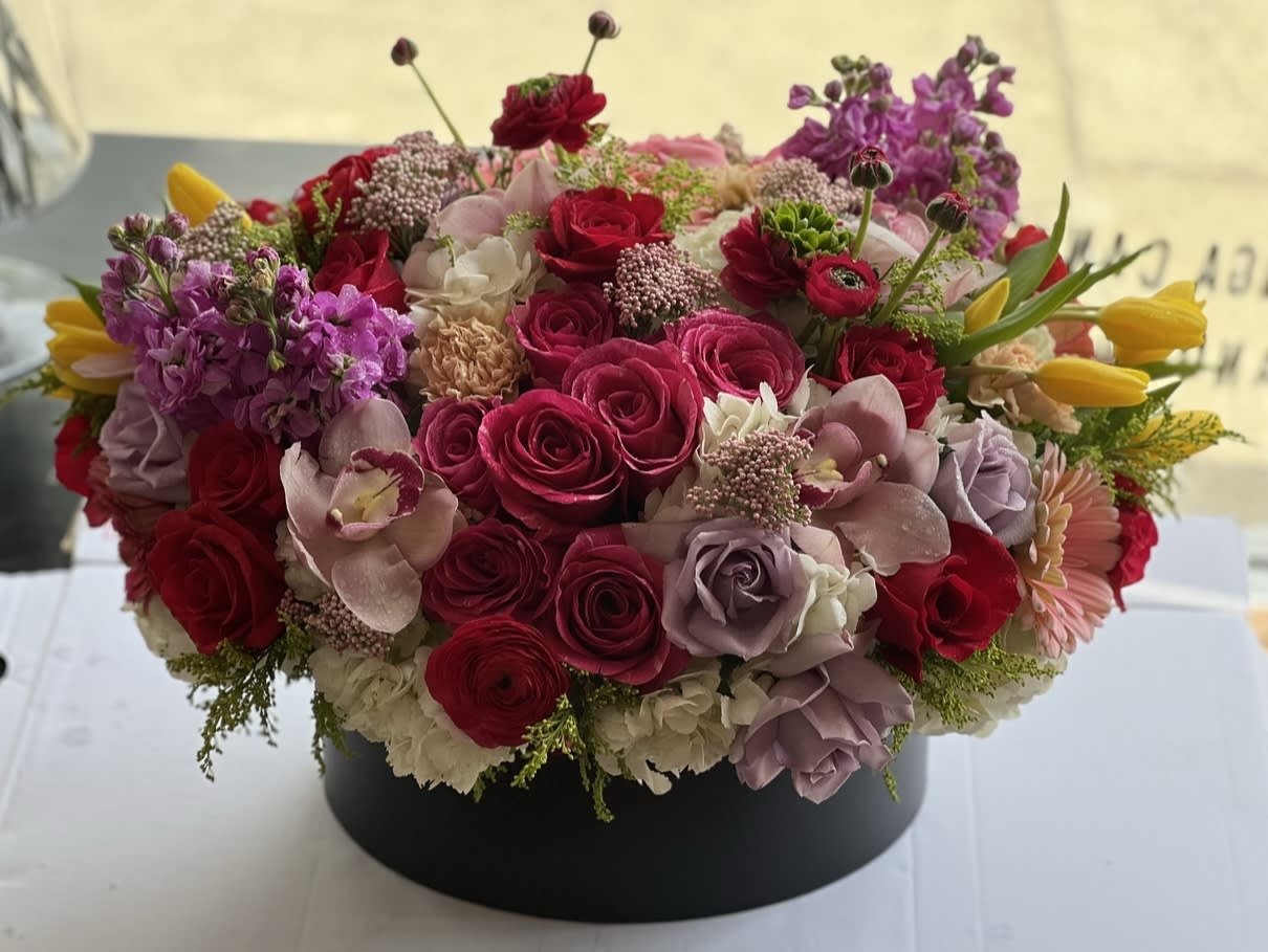 Round arrangement of pink and red roses with purple blooms in a black box