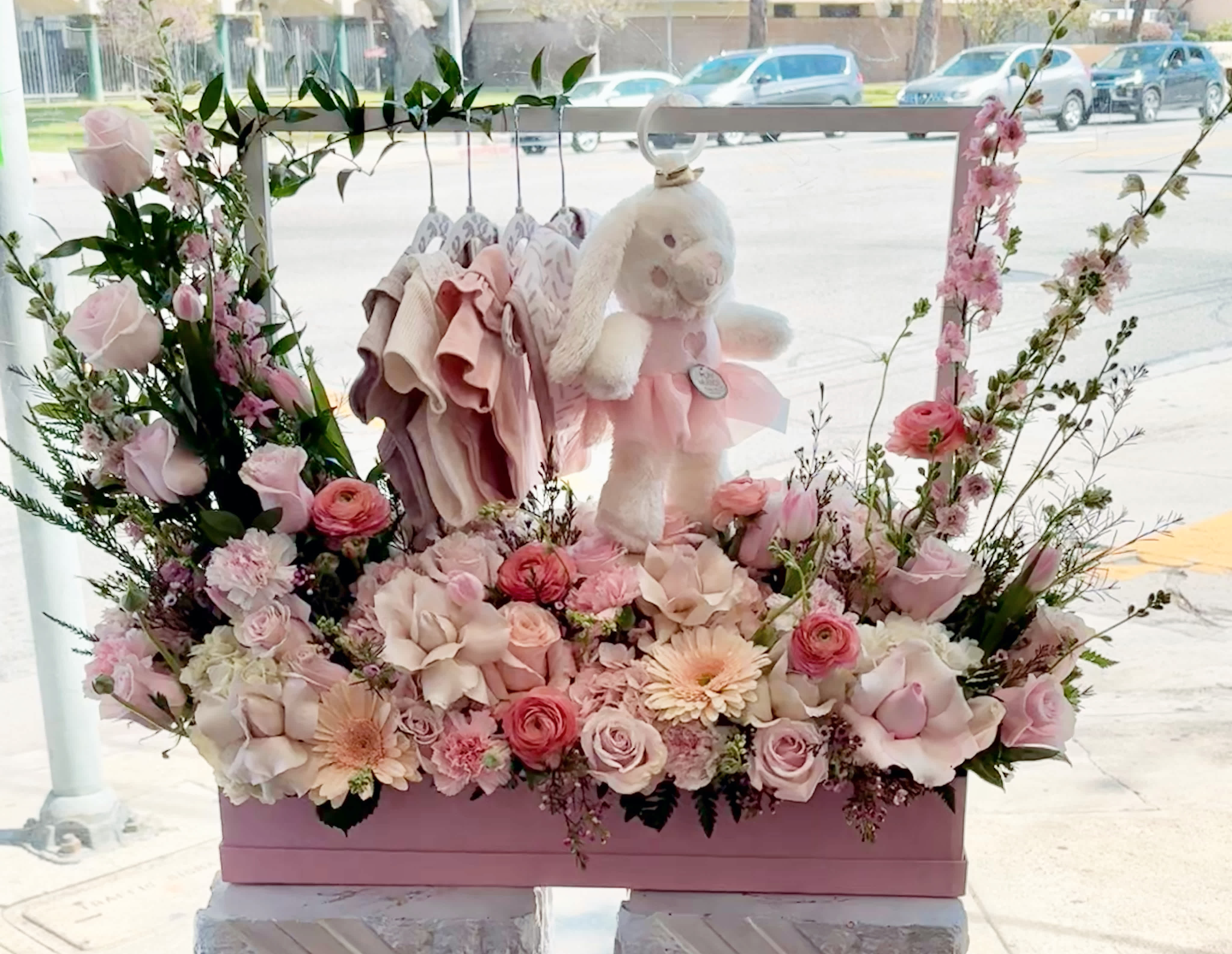 Pink floral display with a teddy bear and baby clothes in a box