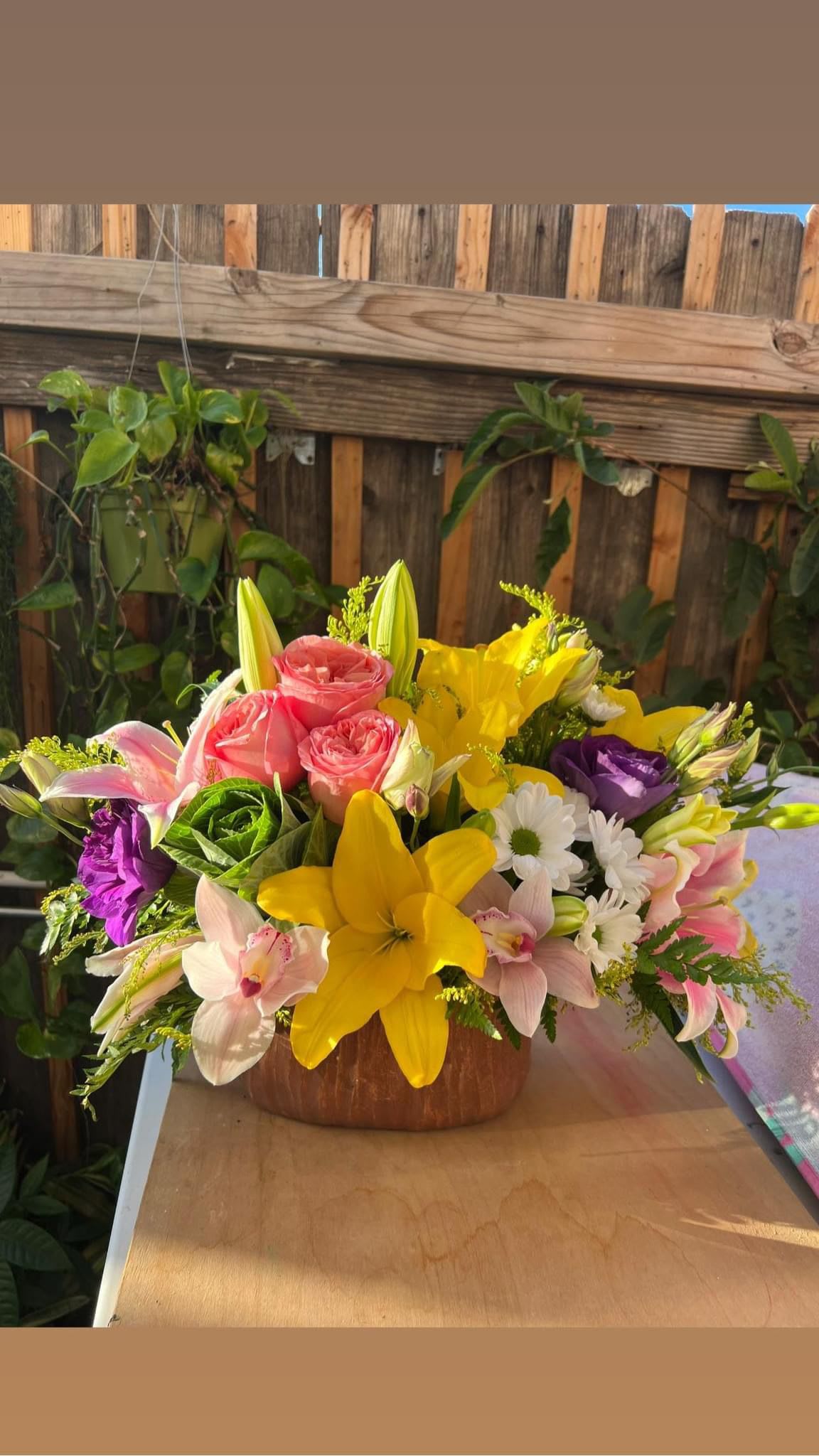 Mixed bouquet with pink roses, yellow lilies, and purple blooms in a wooden bowl