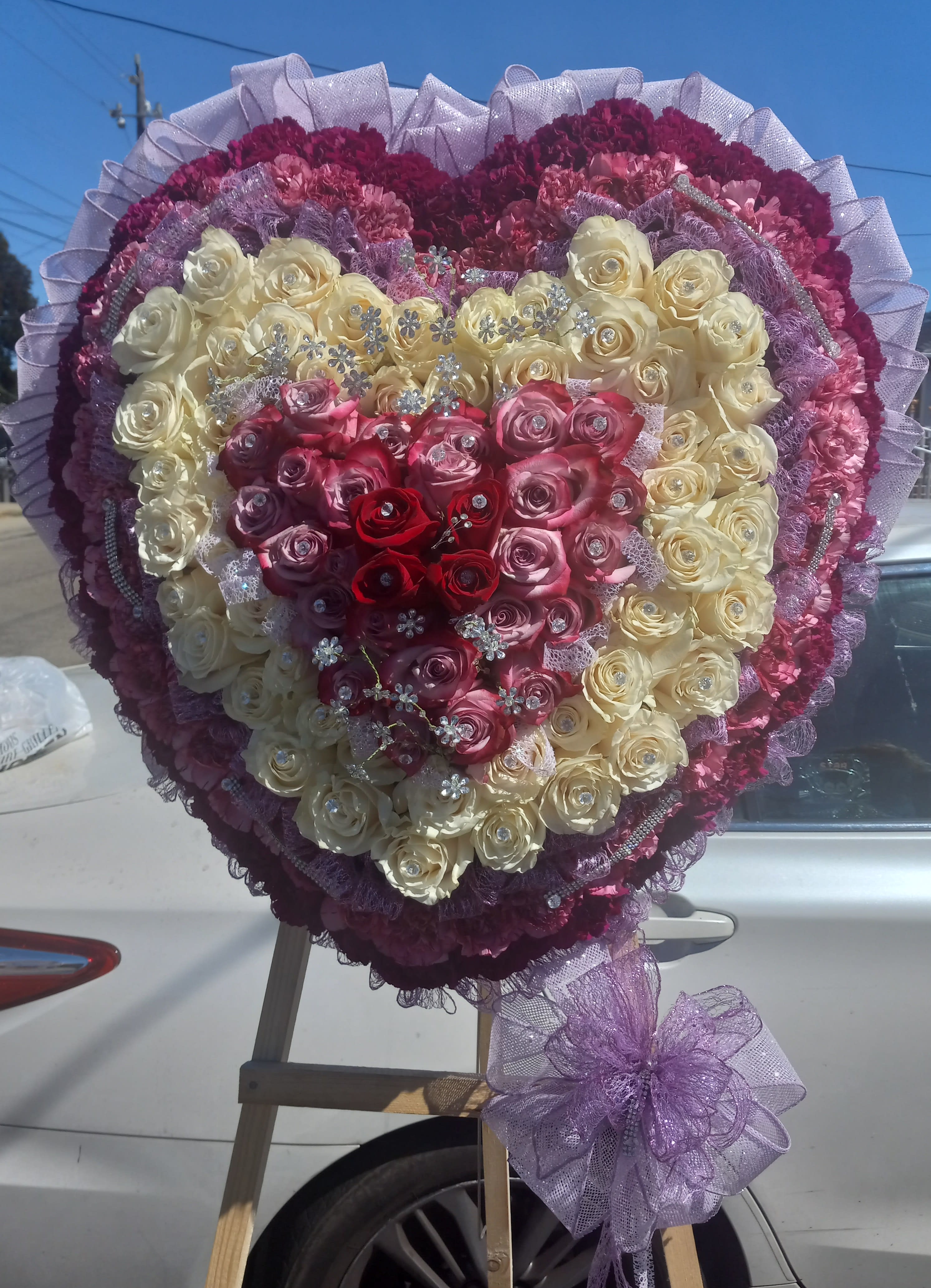 Heart-shaped rose arrangement in cream, pink, and red with purple ribbon trim
