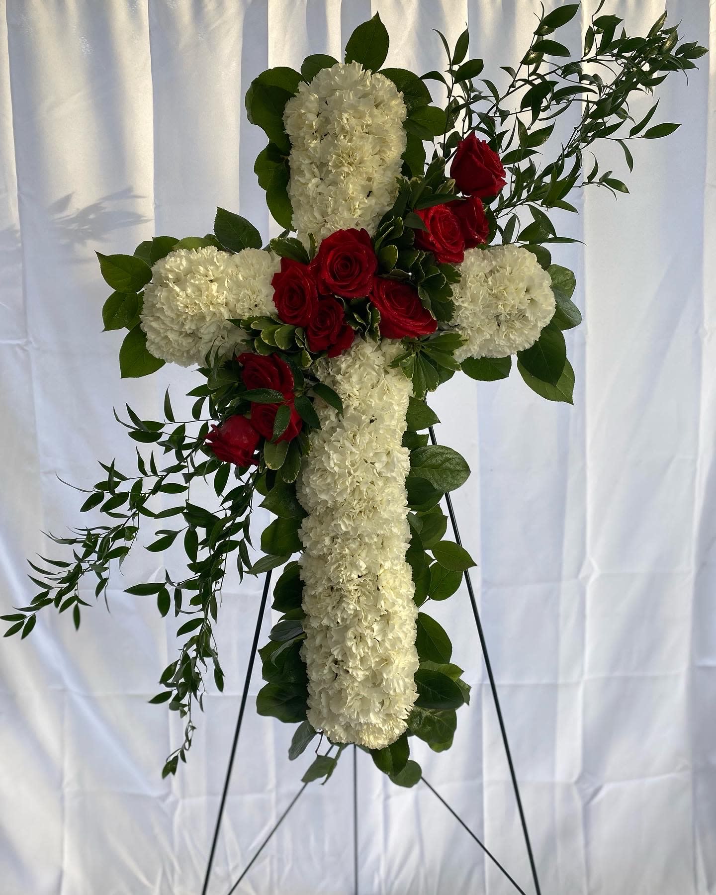 Tall white floral cross with red roses and greenery displayed on a metal easel