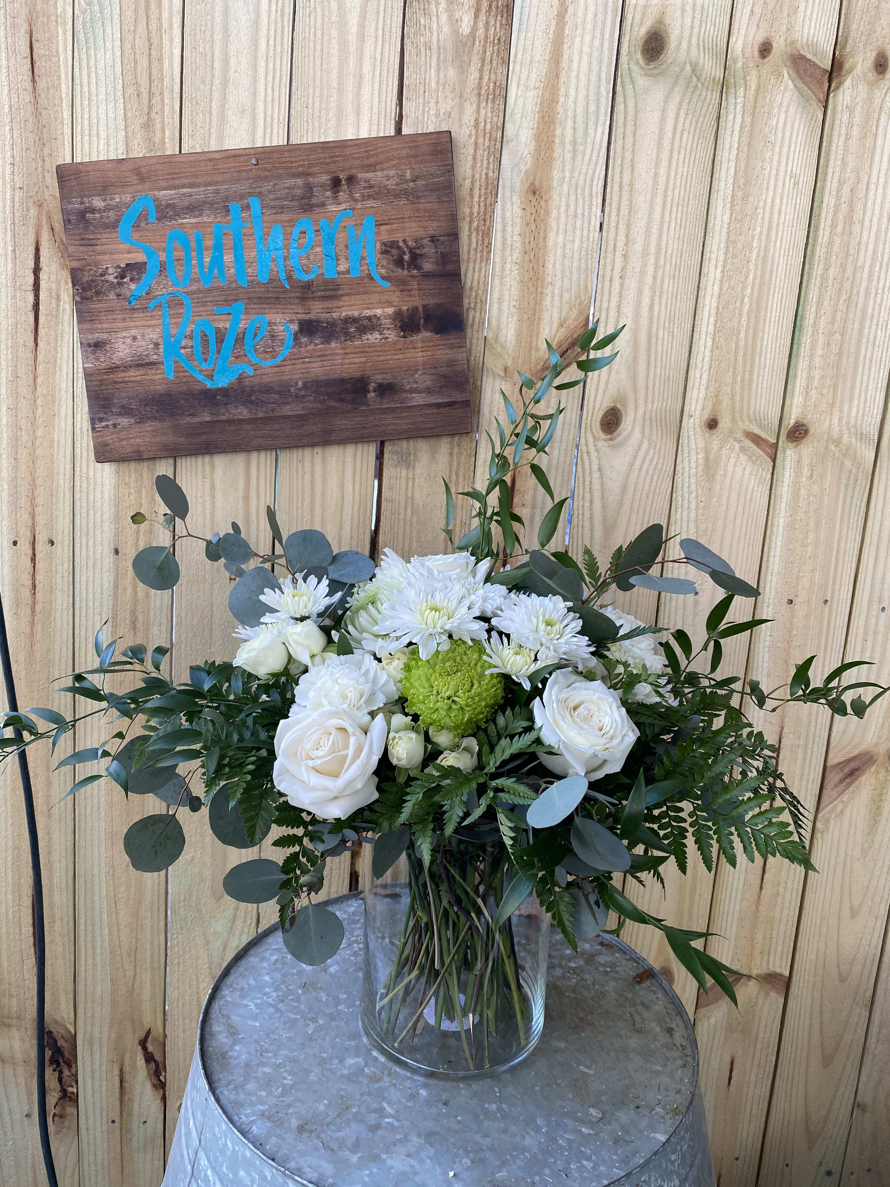 White roses and mums with a green accent bloom in a clear glass vase on a metal table.