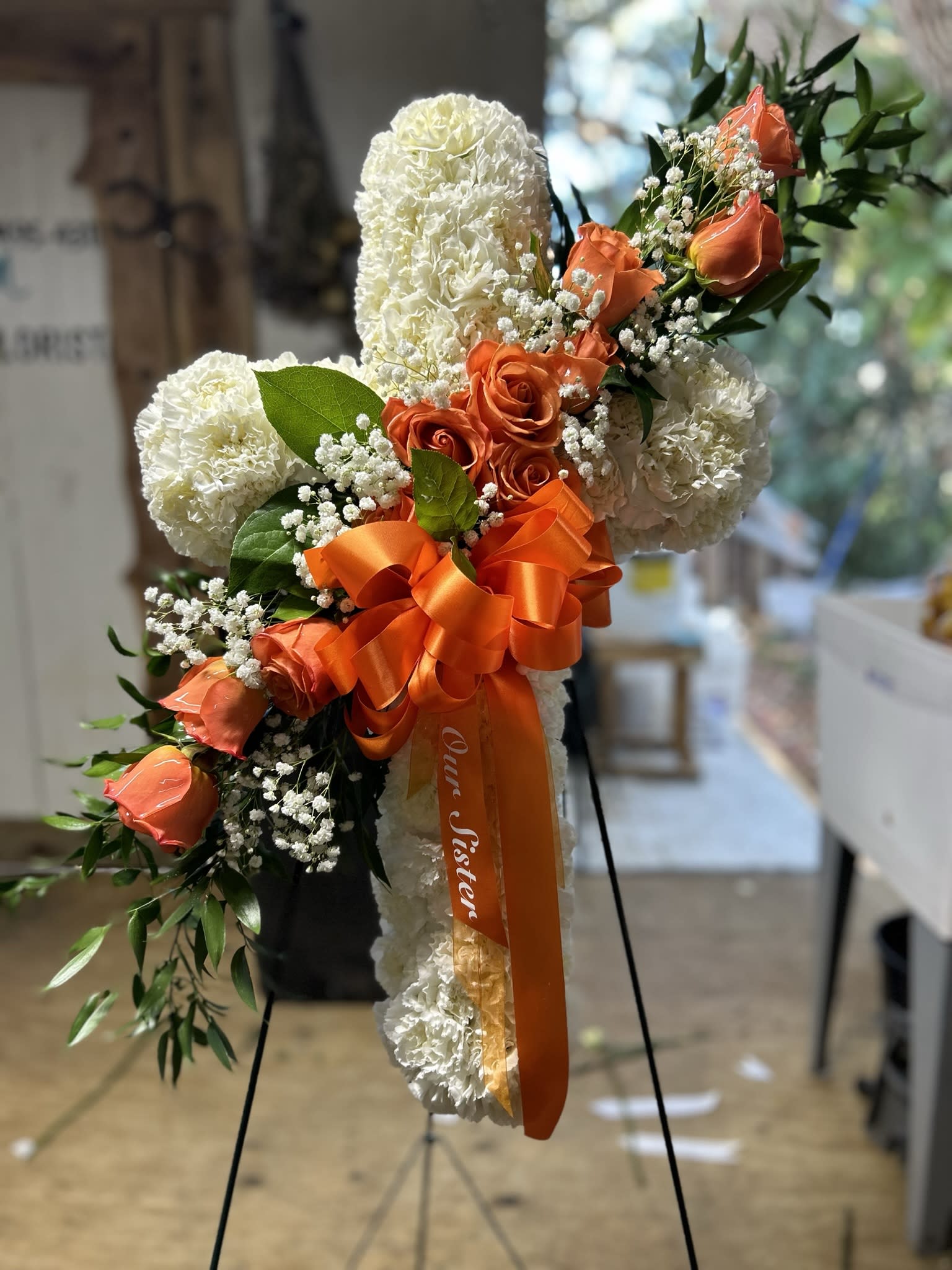 Standing white floral cross with orange roses, ribbon, and greenery on a metal easel.