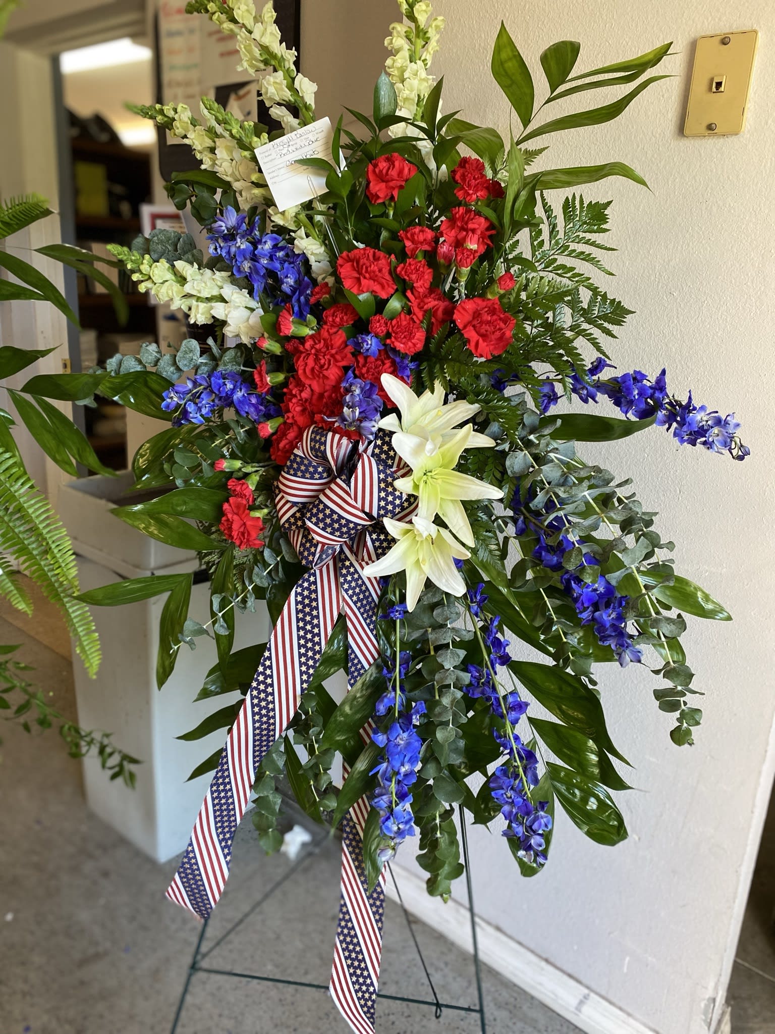 Patriotic standing spray with red carnations, white lilies, and blue blooms on an easel with flag ribbon.