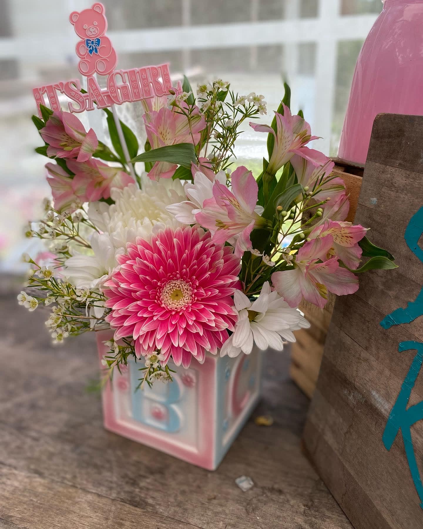 Pink and white baby-themed flower arrangement in a pastel block vase with It's a Girl pick