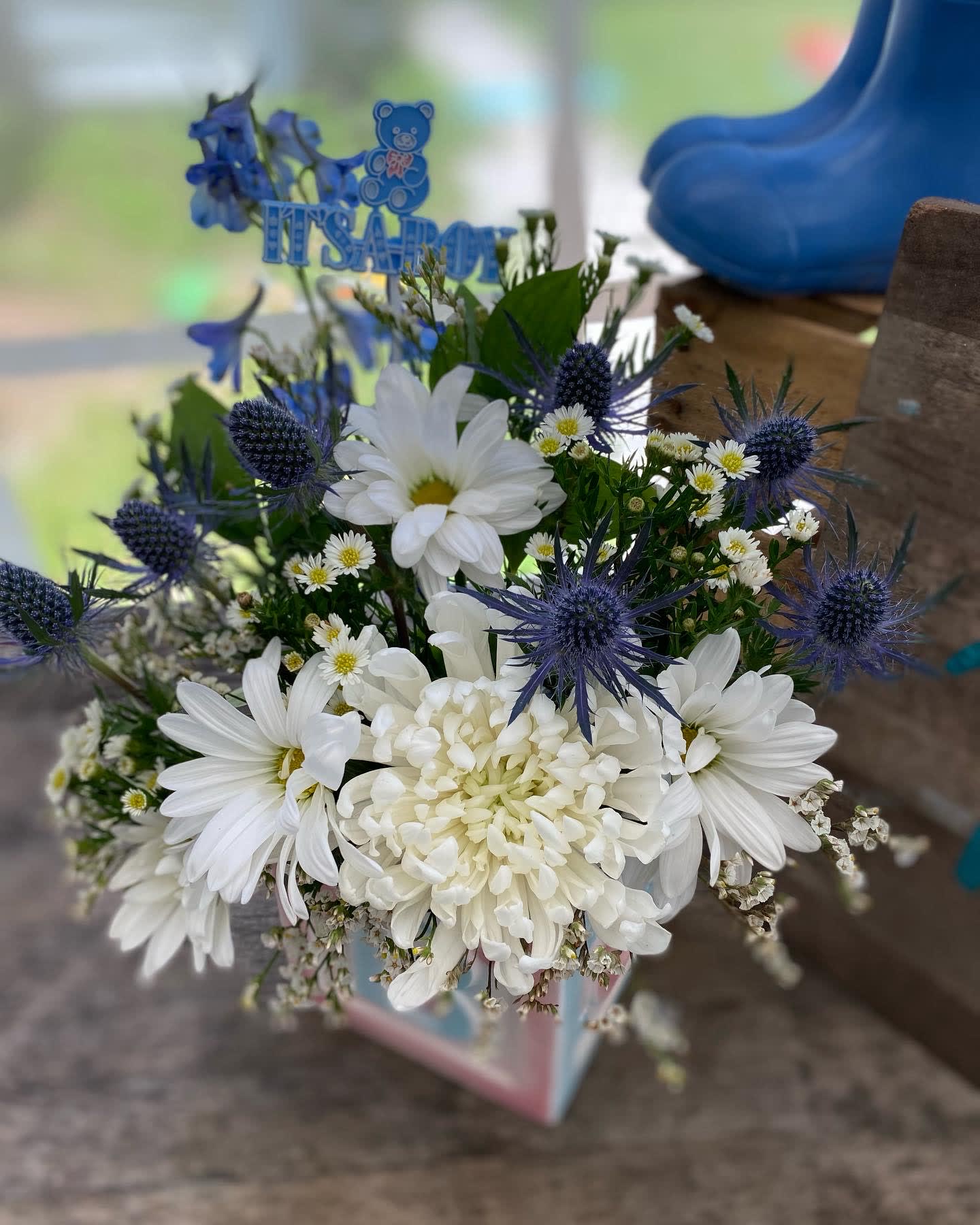 White daisies and mums with blue thistle-like blooms in a cube vase with an 'It's a Boy' pick