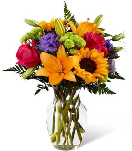 Bright mixed bouquet of lilies, roses, sunflowers, and purple blooms in a glass vase