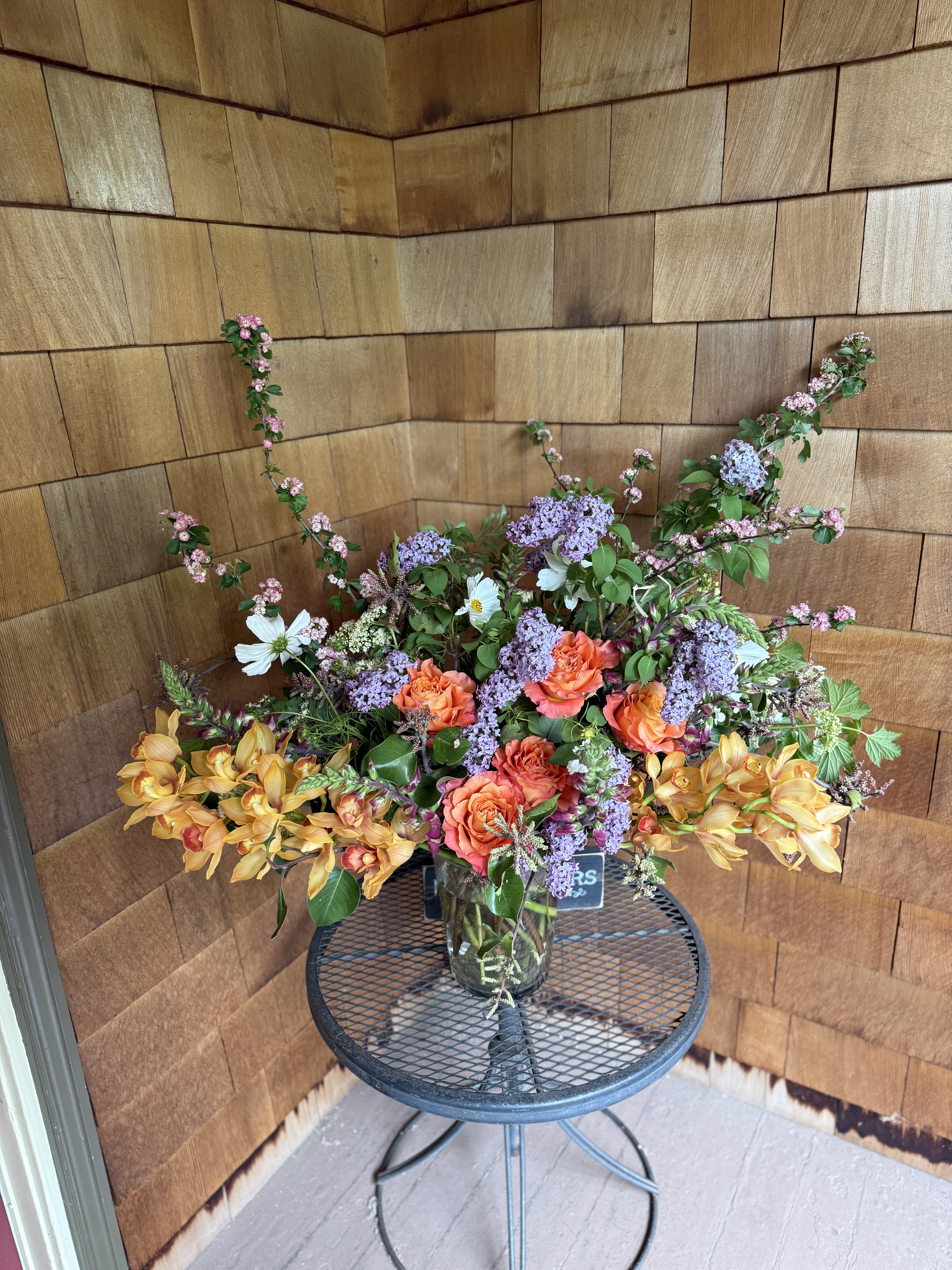 Last supper - A refined expression of natural beauty, this bespoke floral arrangement captures the warmth of a late-summer garden in full bloom. Featuring lush peach garden roses, delicate lavender clusters, and cascading sprays of golden orchids, the composition is artfully balanced with fresh greenery and airy wildflower accents.  Designed in a clear glass vessel, each stem is thoughtfully placed to create movement, depth, and an effortless sense of abundance. The soft interplay of warm apricot, blush, and lilac tones evokes both romance and sophistication, making it a striking centerpiece for any curated space.  Perfect for elevating interiors, special occasions, or gifting with intention, this arrangement embodies timeless elegance with a modern, organic touch