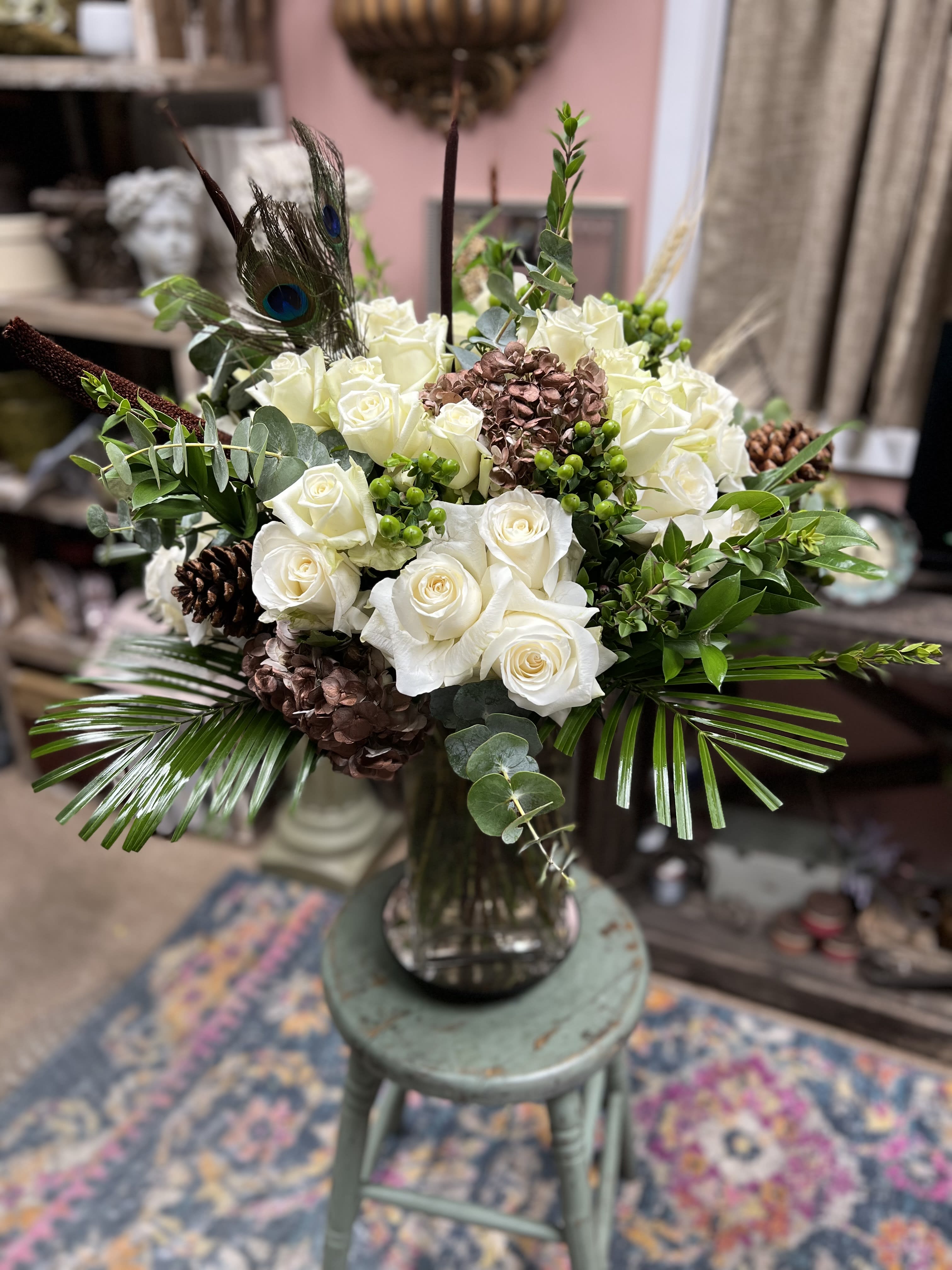 White roses arranged in a glass vase with peacock feathers and pinecones