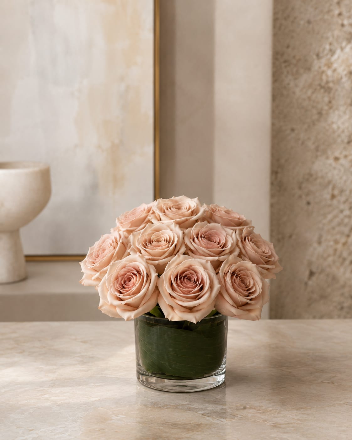 Touch of quicksand  - A compact dome of blush and pale peach roses creates a soft, romantic look-perfect as an anniversary bouquet, wedding table centerpiece, or a refined everyday gift to show someone they're cherished. This flower arrangement places the rose-filled dome front and center so the recipient will immediately feel noticed and celebrated by the delicate layers of petals and whisper-soft pinks.  Tightly gathered roses sit in a clear cylindrical glass vase with dark green foliage at the base to give the design a grounded, polished finish. The neutral, restrained palette of pink, white, and green keeps the silhouette calm and elegant while the compact size makes it ideal for tabletops, bedside tables, or a desk. Simple care: keep water fresh and trim stems as needed to extend bloom life.