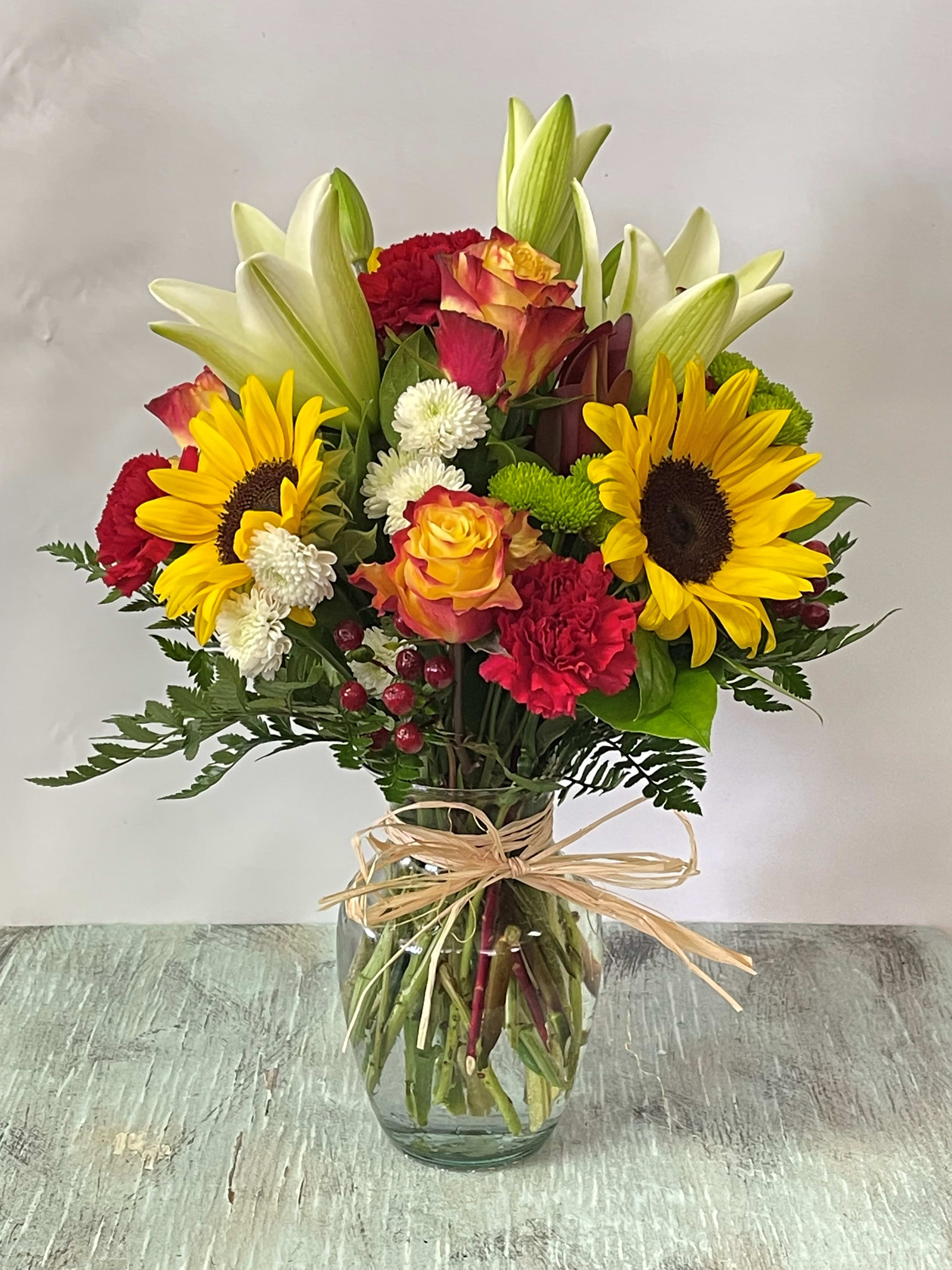 Mixed bouquet of sunflowers, roses, lilies, and carnations in a glass vase
