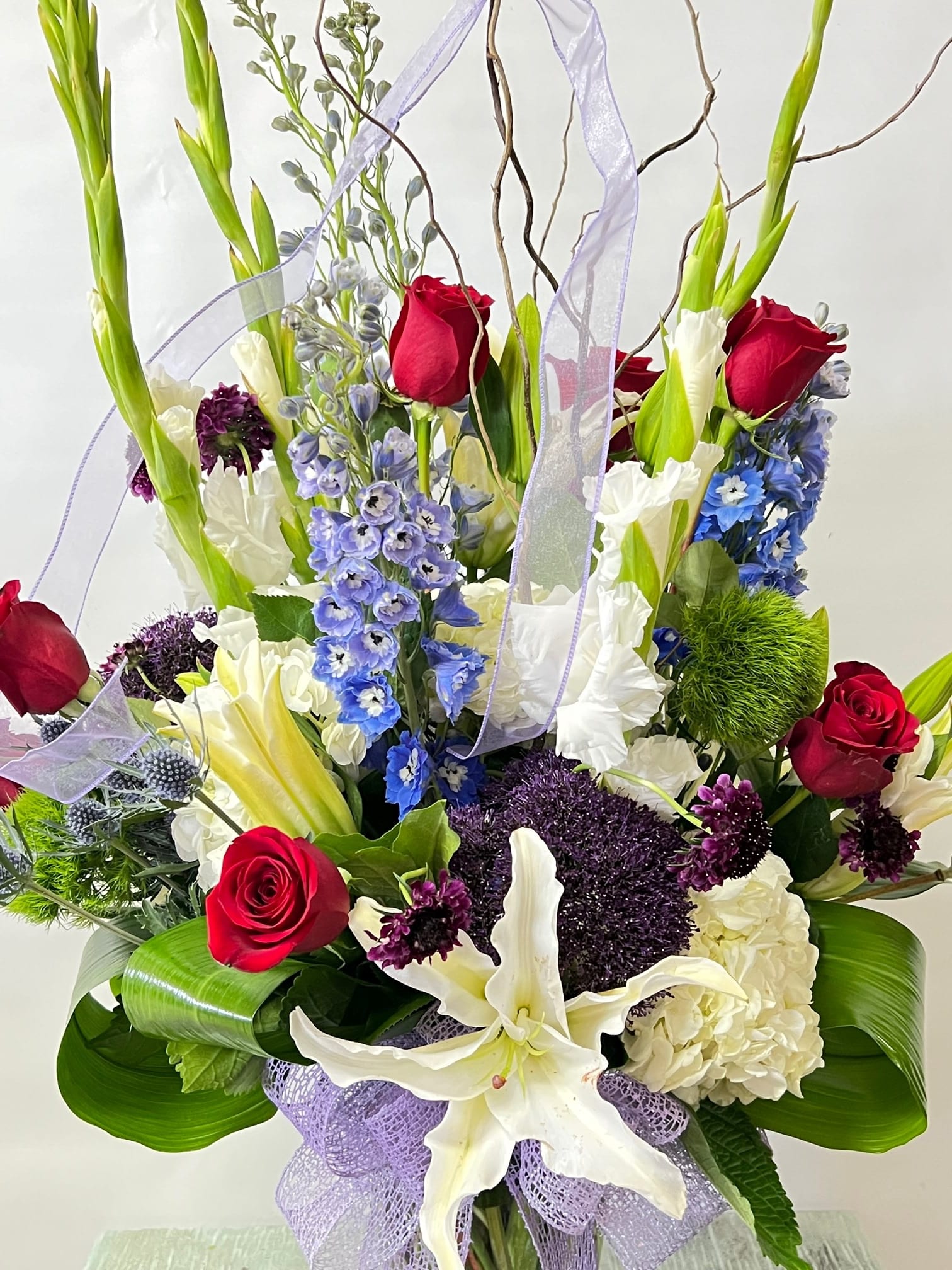 Tall bouquet of red roses, white lilies, and blue flowers with lavender ribbon