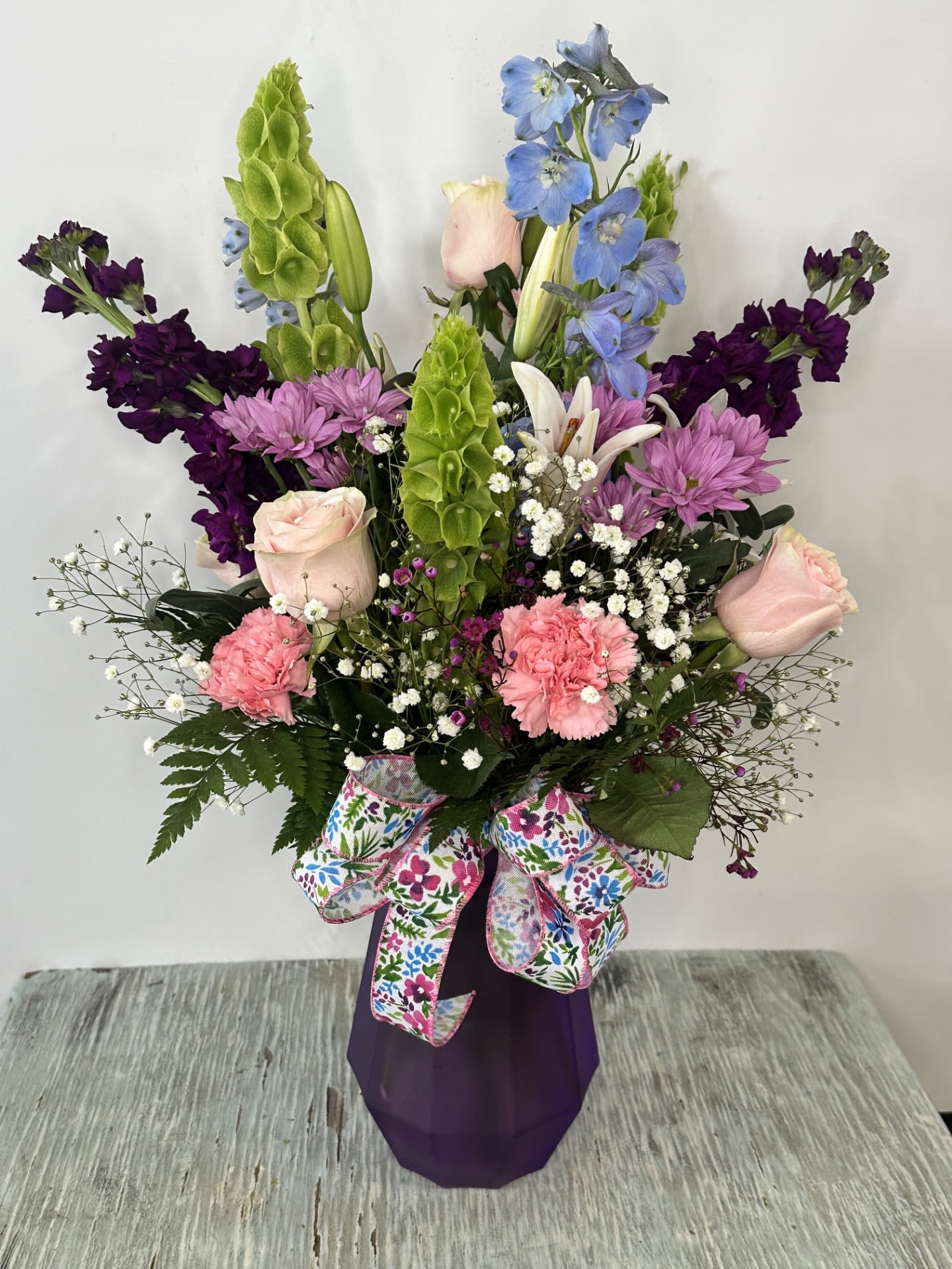 Tall mixed bouquet with pink roses, purple blooms, and blue flowers in a purple vase