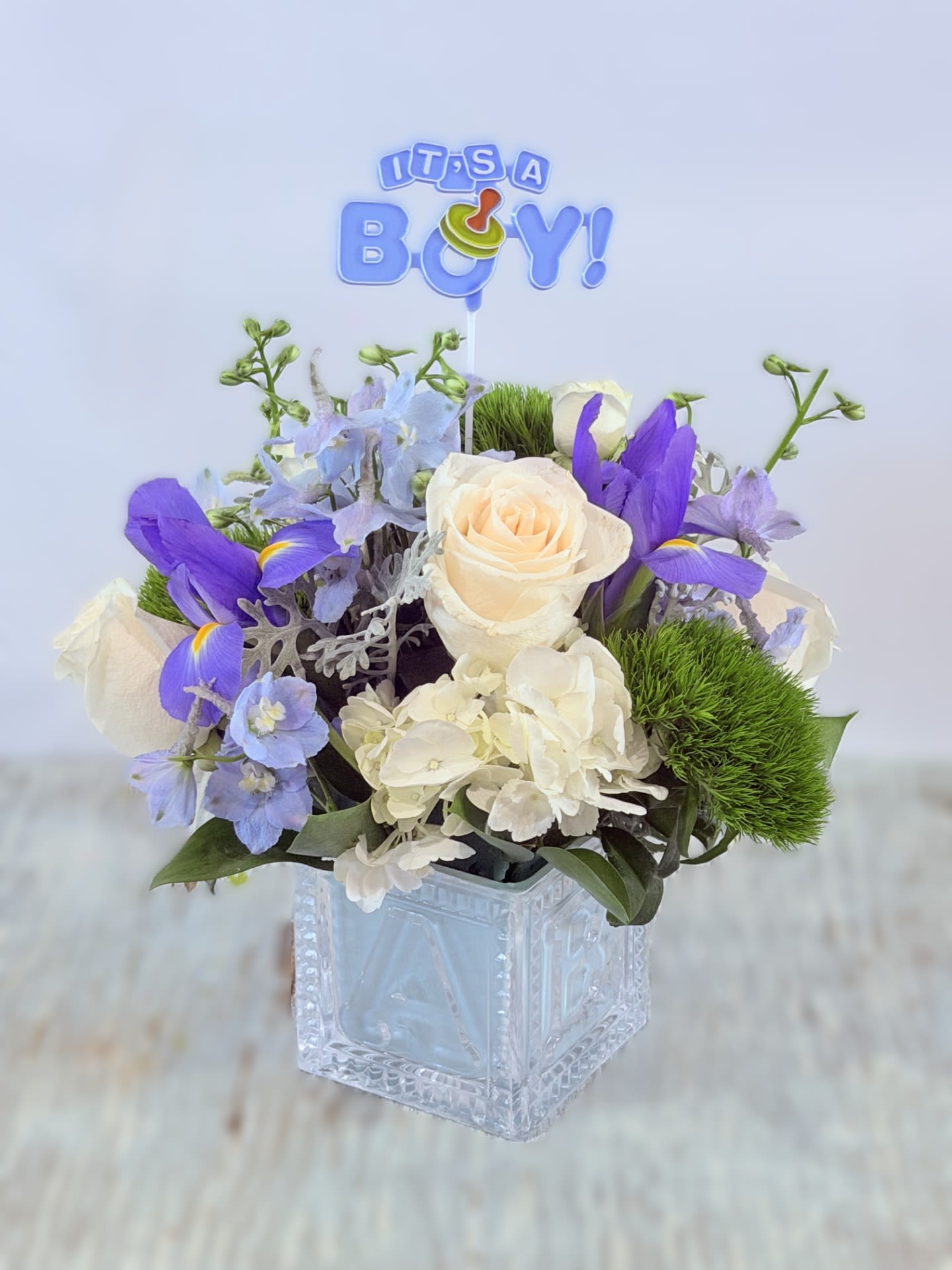 Welcome Baby Boy - Cream roses and cloud-like white hydrangea meet vivid violet and pale blue blooms in a compact, celebratory arrangement made for greeting a newborn - perfect as a baby shower centerpiece or a welcome baby boy gift. The "Welcome Baby Boy" theme is clear and cheerful with a small "It's a Boy!" topper, so the person you're honoring will instantly know this bouquet marks a joyful arrival.  This floral arrangement features cream roses, white hydrangeas, blue delphinium and pale blue iris woven with touches of green foliage for fresh contrast, all arranged in a clear square glass vase. The design is full and rounded with a polished, gift-ready presentation that reads both elegant and playful; the low square vase keeps it easy to display on a dining table or nursery dresser.  Send this to new parents to celebrate a first cuddle, a big brother or sister announcement, or to brighten a hospital room - she'll feel truly celebrated by the soft palette and thoughtful topper. The compact size makes it simple to place and enjoy; no special care beyond fresh water and a cool spot will keep the blooms looking their best.