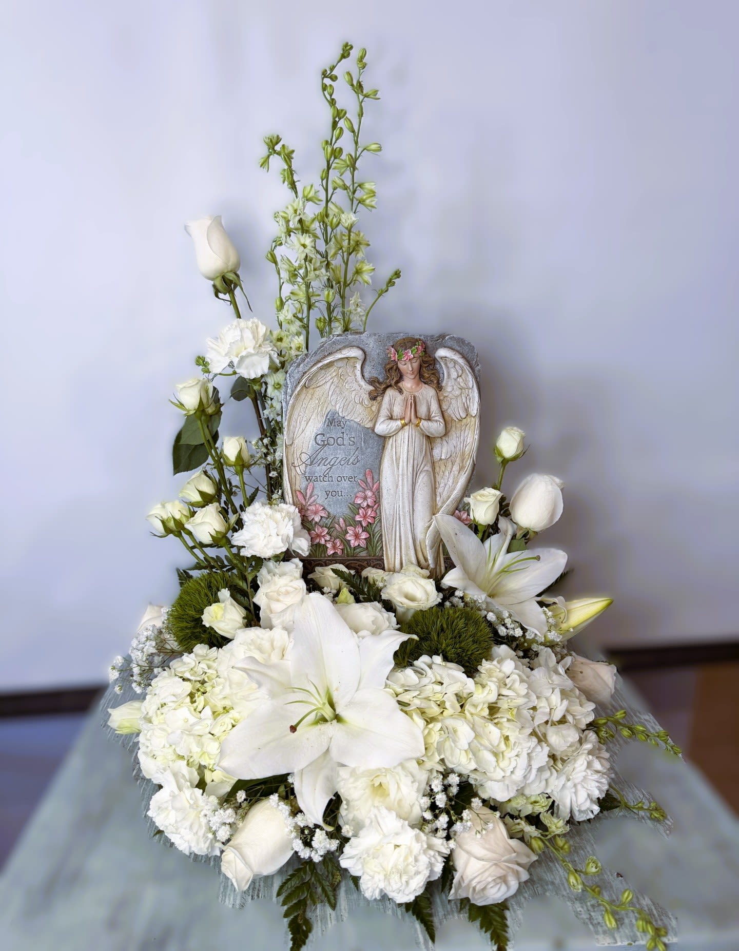 Sympathy #22 - A devotional sympathy arrangement centered around an angel plaque, designed as a memorial tribute to comfort grieving loved ones. This sympathy arrangement features a soft palette of white, green, and a touch of pink and reads as a formal funeral or remembrance piece meant to convey peace and reverence to the person you're honoring.  Large white lilies and white roses take the lead, complemented by white carnations and clustered hydrangea-like blooms, with tall green accent stems adding vertical presence and a layered, full silhouette. A decorative angel plaque with the words "May God's Angels watch over you" and a praying figure is included as a tangible keepsake, and the overall texture is lush yet serene-quiet blooms and gentle greenery that soothe the eye.  Choose this tribute for a funeral service, graveside remembrance, or to send condolences to someone in mourning; it speaks of comfort, faith, and thoughtful remembrance. This design is intended as a formal sympathy piece-please indicate delivery date and any service details when ordering so we can place the arrangement appropriately.