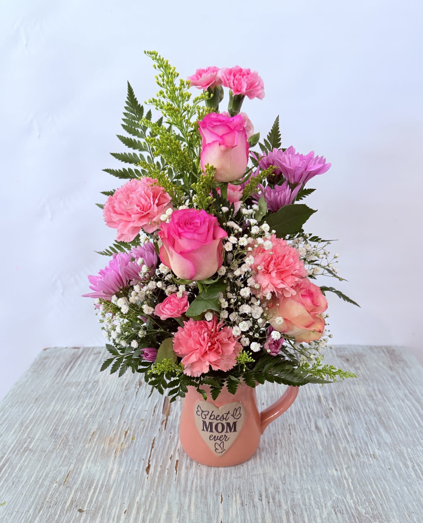 Mother's Day # 2 - A soft-centered pink rose and ruffled carnations set a tender tone in this Mother's Day flower arrangement, making it the kind of gift that makes mom feel genuinely seen and celebrated. Designed as a birthday, Mother's Day, or "just because" gift for a mother or mother-figure, the compact, feminine bouquet pairs blush and lavender tones with fresh white blooms to create a warm, affectionate impression.  This arrangement includes pink roses, pink carnations, white ranunculus, lavender chrysanthemums, alstroemeria, and complementary greenery arranged in a rounded, layered shape. It comes styled in a white 16oz ceramic pitcher/mug decorated with a floral illustration and the word "Mom" (the mug also appears in a peach-pink variation reading "best MOM ever" in some pictured styles), and the finished design measures approximately 11"H x 8"W. Each piece is individually gift boxed; flowers may be substituted as needed to maintain the overall color palette and style while keeping the same sentimental look.