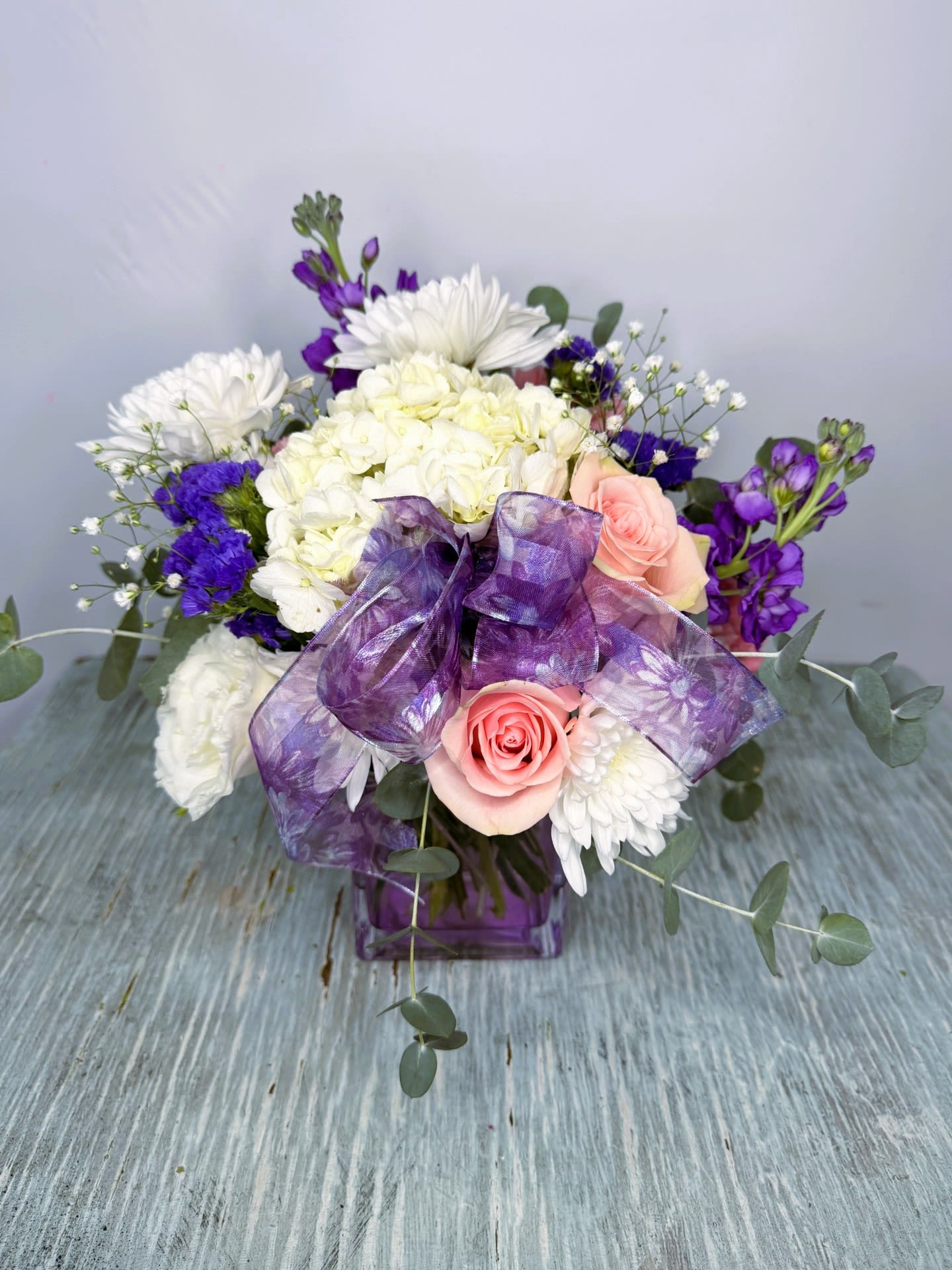 Mother's Day #4 - A compact, rounded Mother's Day flower arrangement that feels fresh and romantic-perfect as a birthday bouquet, thank-you gift, or springtime surprise for the special woman in your life. White chrysanthemums and pale pink roses create a soft, tender core while vivid purple filler blooms add lively contrast; the design reads both gentle and slightly dramatic, the kind of bouquet that makes her feel truly seen.  Arranged in a purple glass vase and embellished with a sheer purple ribbon, this piece mixes texture and airiness: eucalyptus-like greenery frames the blooms and gives the arrangement a loose, natural outline. The color story-white, pink, purple, and green-works beautifully for mothers, friends, or mentors who love classic florals with a modern twist. Compact enough for a side table yet full of presence, it arrives ready to display; simple care-fresh water and a cool spot away from direct sun-will keep the flowers looking their best.