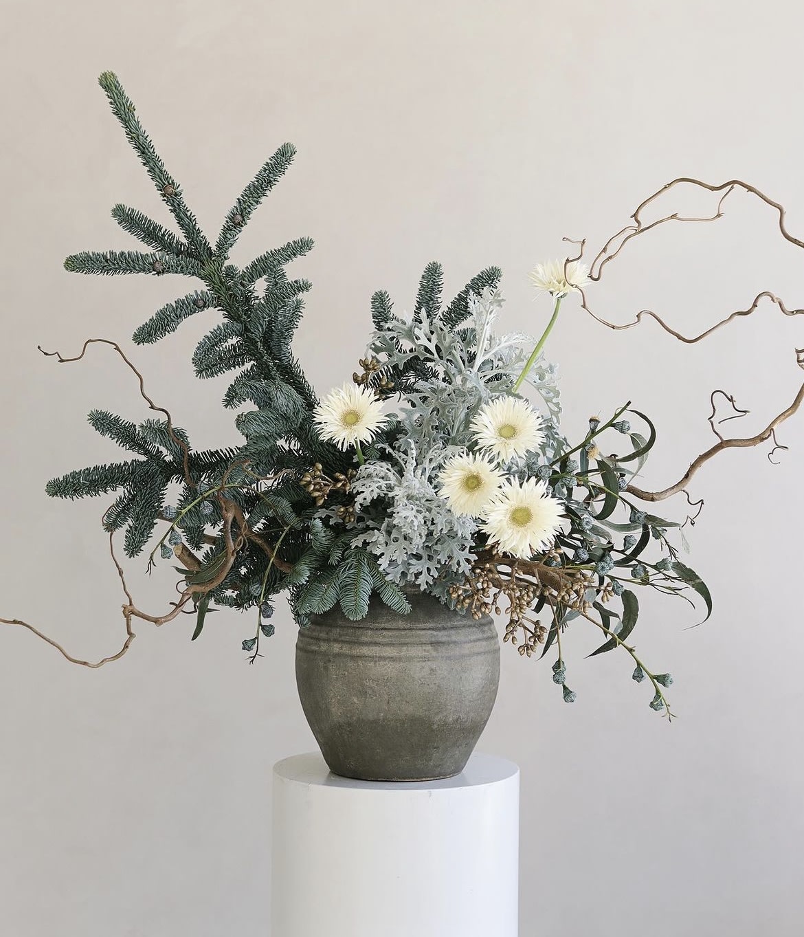 White daisy-like flowers in a gray vase with evergreen branches