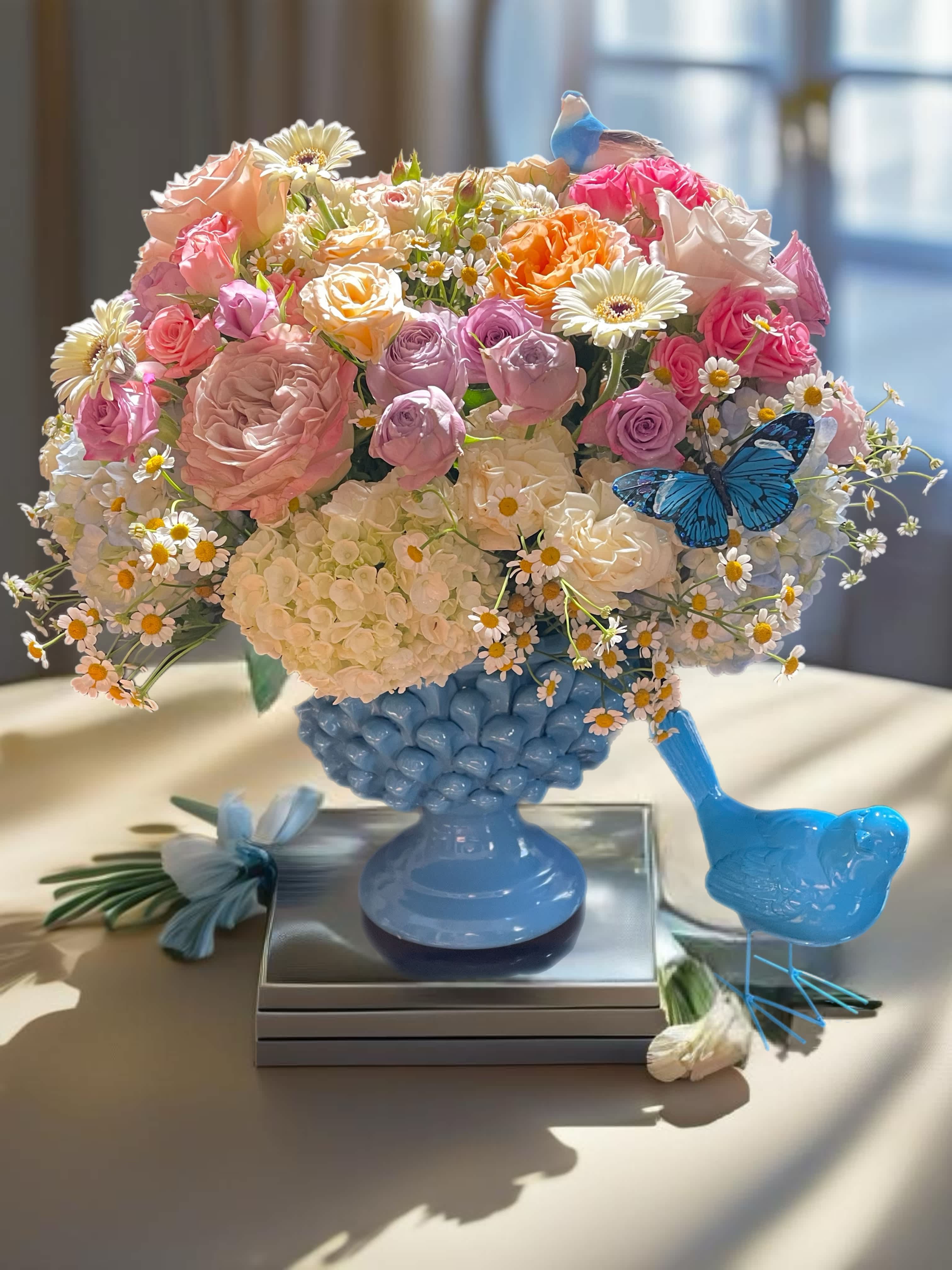 Pastel bouquet of roses, hydrangeas, and daisies in a blue vase with bird accents
