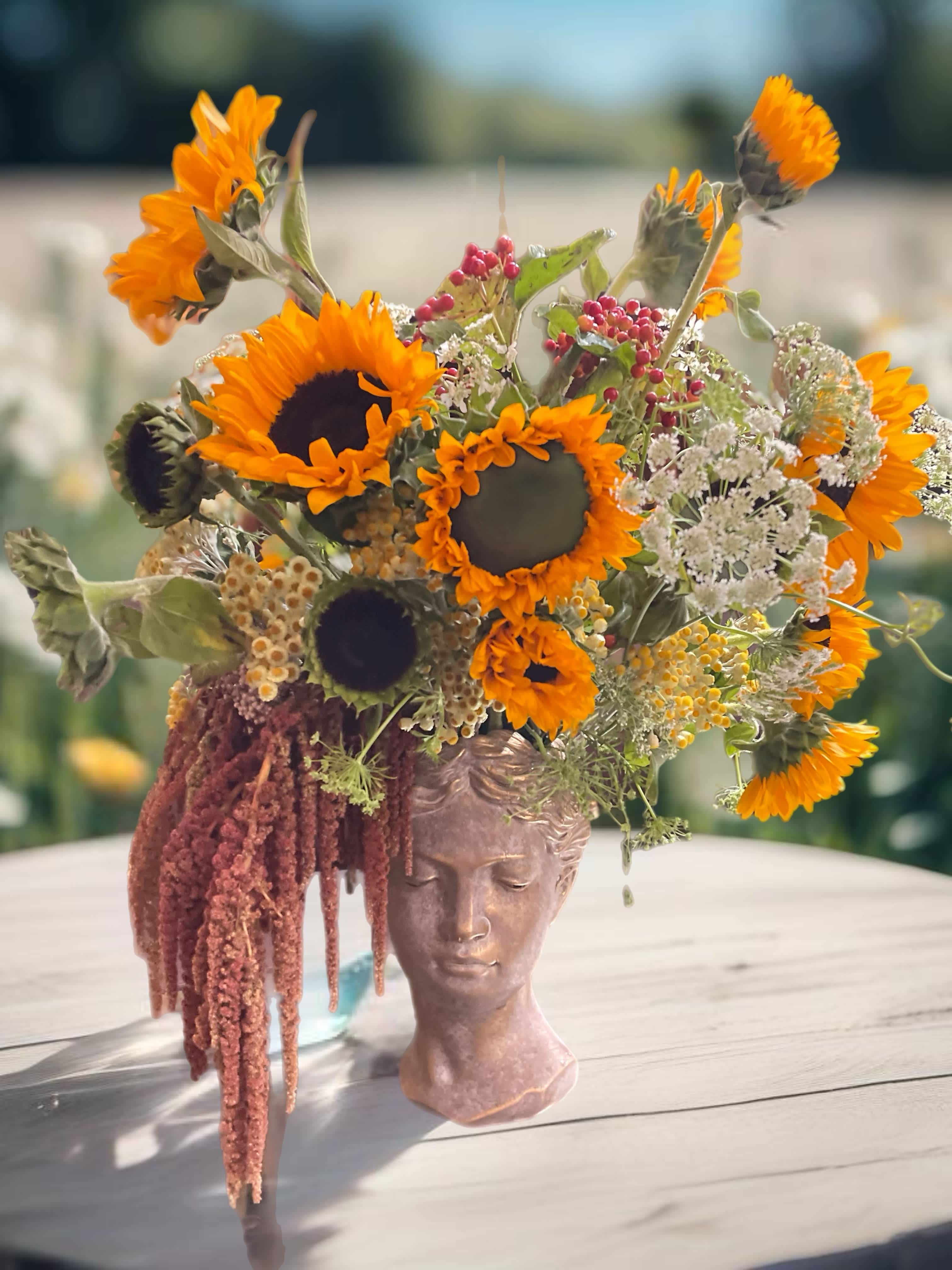 Sunflower arrangement in a sculpted face vase with trailing burgundy amaranthus