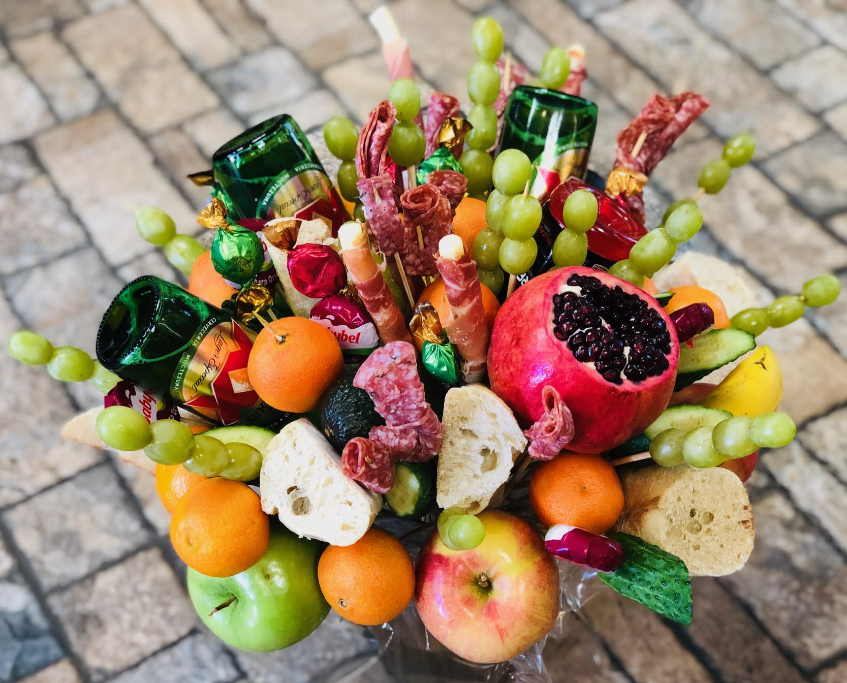 Boozy Gourmet   - Bright, abundant, and unmistakably festive, this Boozy Gourmet edible bouquet is a celebration in a round container-perfect as a birthday bouquet alternative, anniversary gift, or a just-because surprise for the person who prefers savory over sweets. The arrangement reads like a party on a stem: green glass beer bottles angled out for drama, halved pomegranate and bright apples for vivid reds, oranges and grapes for pops of orange and green, plus wrapped candies that add cheerful accents. It's an ideal holiday or Christmas centerpiece, a thoughtful get-well or graduation treat, and a playful way to say "thank you" or "I'm sorry."  This upright display combines fresh fruits, small loaves and rustic bread slices, and rolled salami and prosciutto on wooden skewers for texture and flavor-seasoned, savory, and ready to nibble. Clear cellophane wrap keeps everything secure, and cured meat skewers, wrapped candies, and assorted fresh fruits are arranged densely so each biteable element is easy to reach. Alcohol is not included; the green glass beer bottles are decorative and add to the boozy-gourmet look without shipping restrictions.  