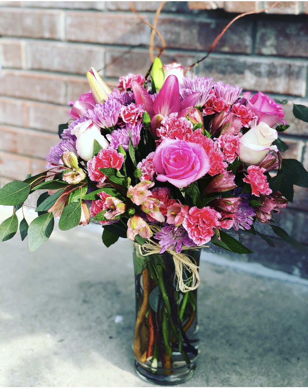 Pink and Lavender Mixed Flower Arrangement in Glass Cylinder Vase - A lush blend of pink roses and soft lavender chrysanthemums creates an instantly romantic birthday or Valentine's Day bouquet that will make the person you're thinking of feel cherished. This pink and lavender mixed flower arrangement in a glass cylinder vase combines feminine pinks, touches of purple, and white highlights for a bright, affectionate mood-perfect for anniversaries, Valentine's Day, or simply to say "you're special."  The arrangement includes roses, carnations, alstroemeria, lilies, and other mixed blooms gathered in a clear glass cylinder vase so the stems show through the water for a fresh, garden-picked look. Finished with a simple raffia ribbon at the neck, the design balances lush texture from hydrangea-like massing and stock with the delicate snapdragon and spray rose accents visible in the photos. Colors range from deep pinks and soft peach to lavender and white, offering a cheery, feminine palette.  Ideal for surprising a partner, brightening a friend's birthday, or sending Valentine's Day wishes, this arrangement reads as both elegant and casually affectionate-the kind of bouquet that makes someone feel seen and celebrated. Available for delivery in Killeen, TX, the vase arrangement arrives ready to display; change the water every few days and trim stems to extend freshness.
