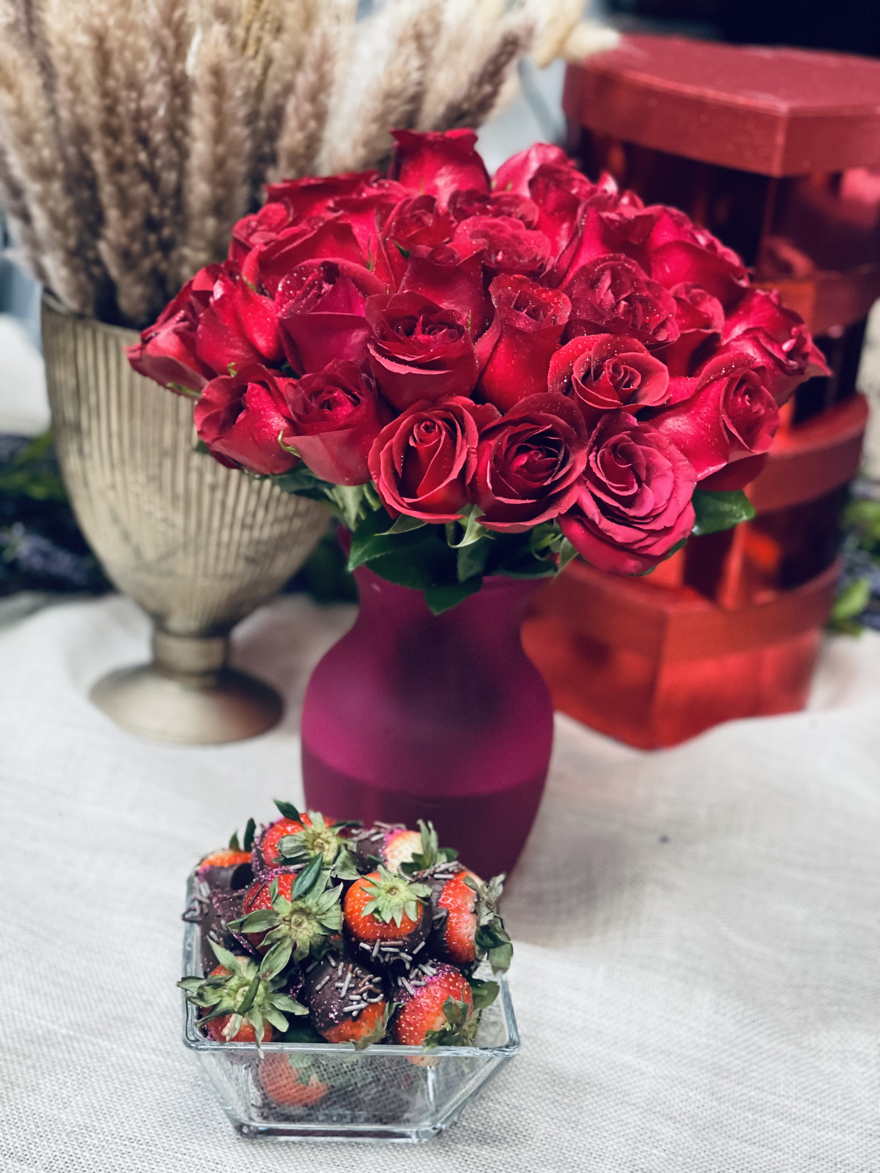Red rose bouquet in a purple vase with chocolate-covered strawberries in front