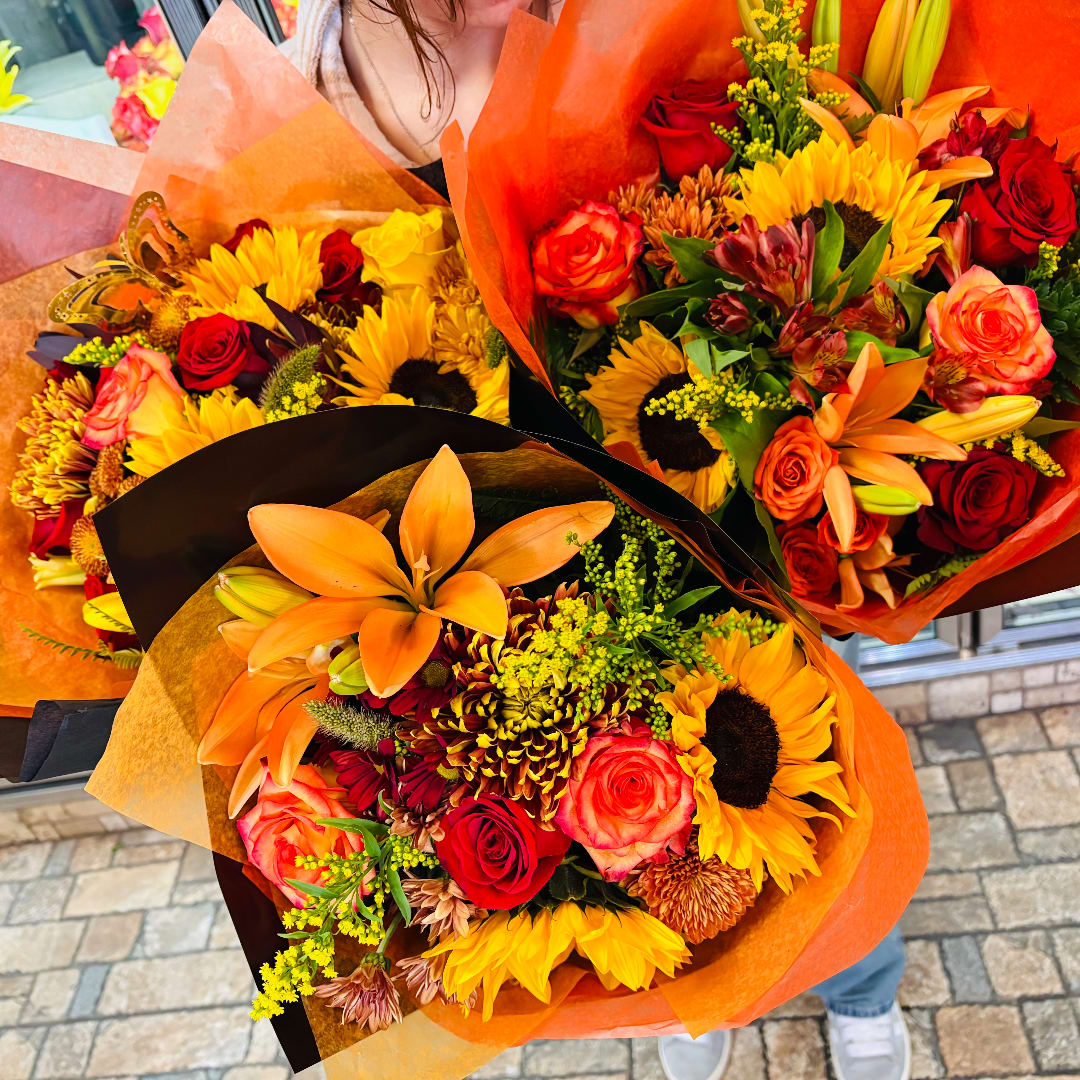 Three colorful bouquets with sunflowers, roses, and lilies wrapped in orange paper