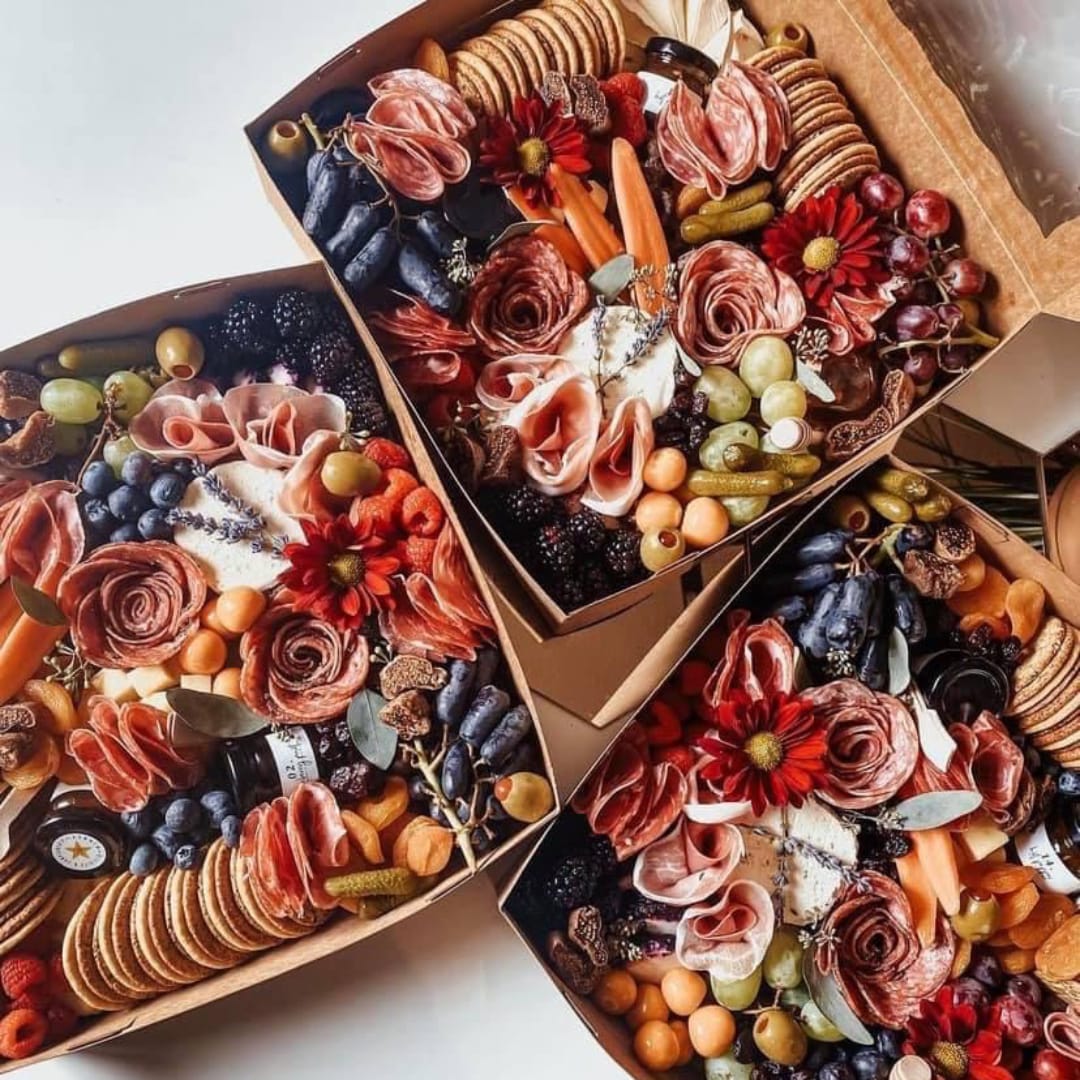 Four charcuterie-style gift boxes filled with fruit, crackers, and flowers
