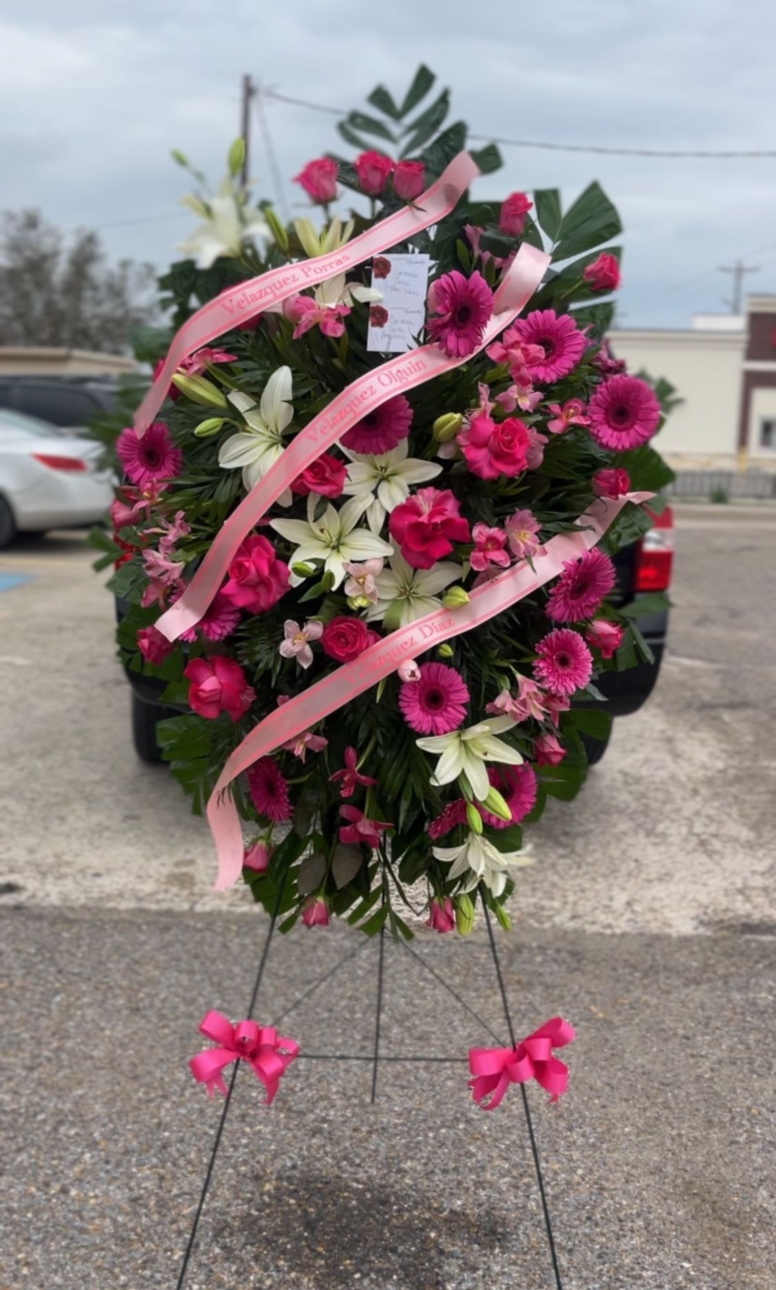pink tribute - Bright, ceremonial pink and white standing spray designed for sympathy and memorials - the kind of funeral arrangement that reads as both respectful and warmly expressive to the person you're honoring. This funeral spray features a dense, oval silhouette of vivid pink gerbera daisies and roses layered with white lilies and delicate pink alstroemeria, arranged to present a strong visual impression from the front of the service. The overall effect is formal and comforting, meant to make mourners feel seen and to convey heartfelt remembrance.  Built on a metal easel with a floral stand, the arrangement is finished with multiple pale pink satin ribbons (some printed with names) that drape across the face for a traditional tribute look. Dark foliage forms a full, contrasting backdrop so the bright pinks and whites pop, and bow accents at the easel base add a composed, ceremonial touch. Included hard goods: standing easel, pink ribbon, floral card, and floral stand.  Choose this piece when you need a prominent, dignified tribute for a funeral, memorial service, or graveside ceremony - especially for someone whose memory you want celebrated with bold, uplifting color. As a large standing spray it's intended for display at services; if you need sizing or delivery specifics, please select your preferred delivery date at checkout or contact us for placement guidance.