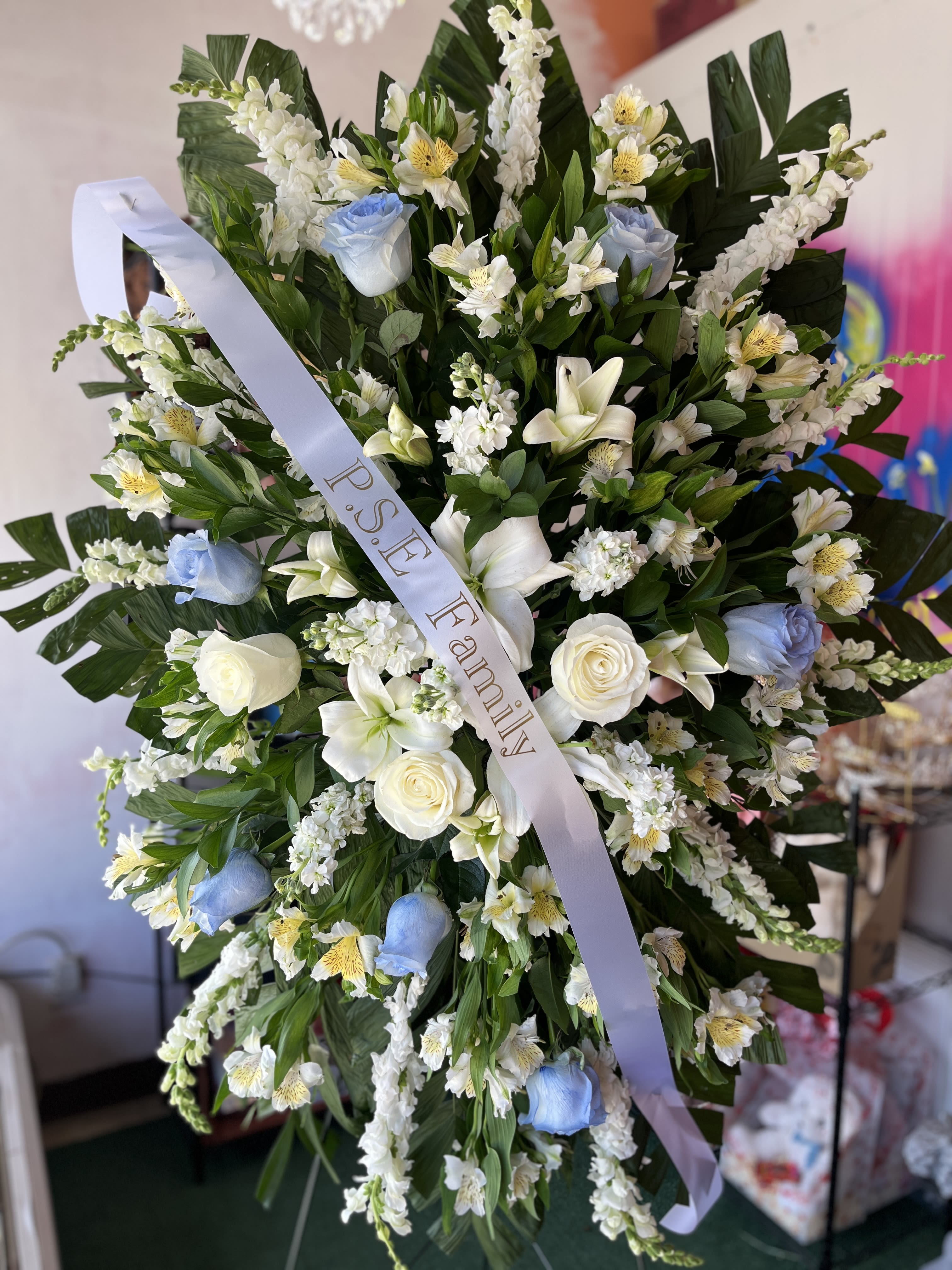 Whisper of Heaven - Soft pale blue roses set against a halo of dense white blooms create a calm, reverent presence-perfect as a sympathy flower arrangement or funeral standing wreath. Whisper of Heaven is designed to comfort the person you're honoring: the restrained palette of white, green, and blue and the formal, symmetrical composition read as a respectful tribute that helps convey sympathy and remembrance.  This large standing wreath features pale blue roses, white lilies, white stock and alstroemeria clustered into a full, layered circle with abundant dark green foliage for depth and contrast. The arrangement is finished with a wide mourning satin ribbon printed "P.S.E Family" across the center, giving the piece a clear memorial tone. Textures range from the smooth rose petals to the trumpet lilies and the delicate filler clusters, producing a composed, dignified look appropriate for a funeral service or graveside display.  Choose Whisper of Heaven when you want the recipient to feel seen and comforted in their loss-a thoughtful, traditional tribute for family members, close friends, or colleagues. As a standing wreath it arrives ready to display at a service; please contact us with service details for delivery and placement instructions.