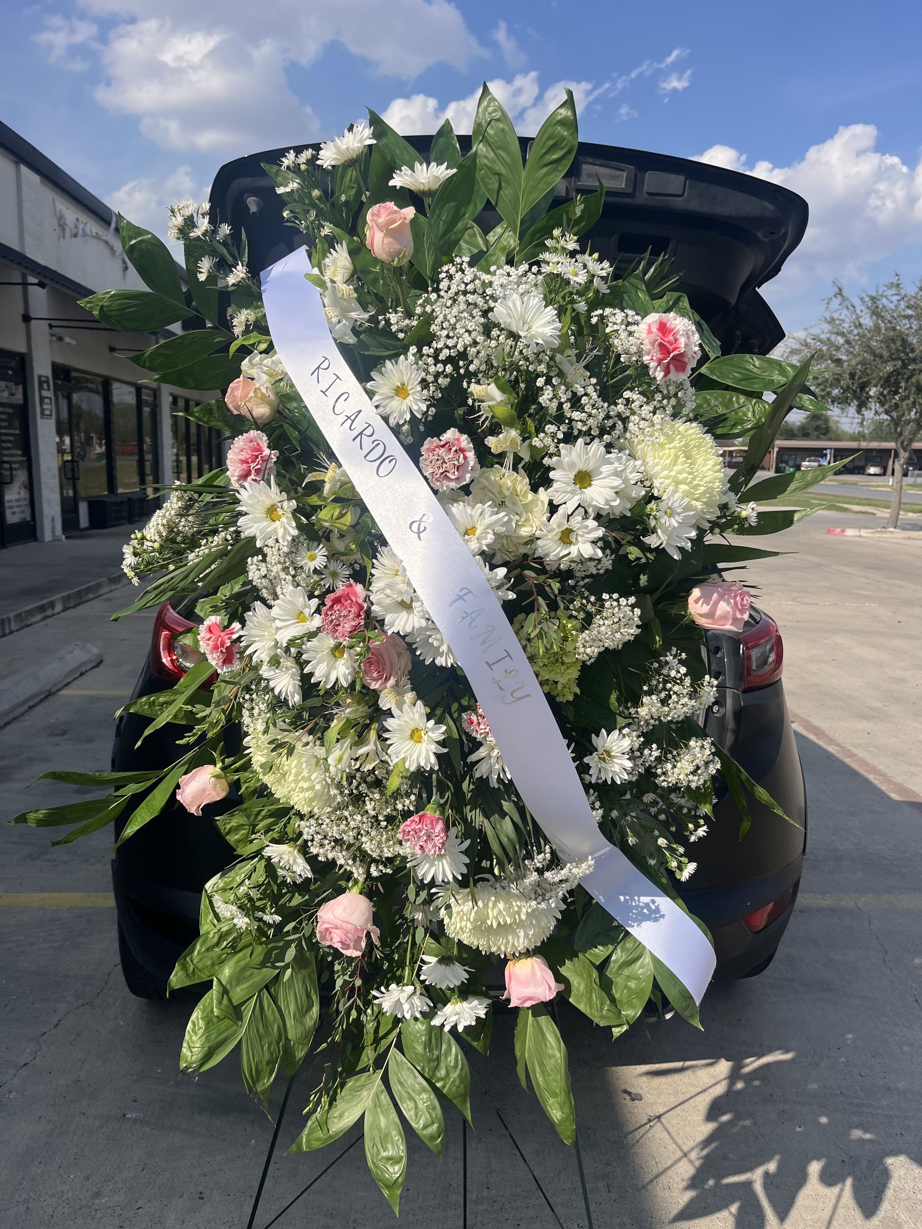 Sweet Repose - A soft, formal standing spray in white, pink, and green designed as a sympathy or funeral tribute - the kind of arrangement that quietly honors a life and comforts the person you're thinking of. The cascading, dense shape reads respectful and abundant; pale pink roses and pink carnations provide gentle color while white daisy-like blooms and clusters of baby's breath keep the palette airy. A white ribbon with printed names across the front gives a clear memorial sentiment suited for a service or graveside display.  This large spray is layered and full, framed by broad glossy foliage that creates a deep green backdrop and a dignified silhouette on a floral stand or standing easel. Textural contrasts - the soft rose petals, ruffled carnations, delicate alstroemeria and tiny filler flowers - make the piece feel handcrafted and substantial without being showy. Sized for prominent display, it arrives ready to place on a stand and includes the white ribbon; it offers a gentle, heartfelt way to say you're thinking of the family during a difficult time.