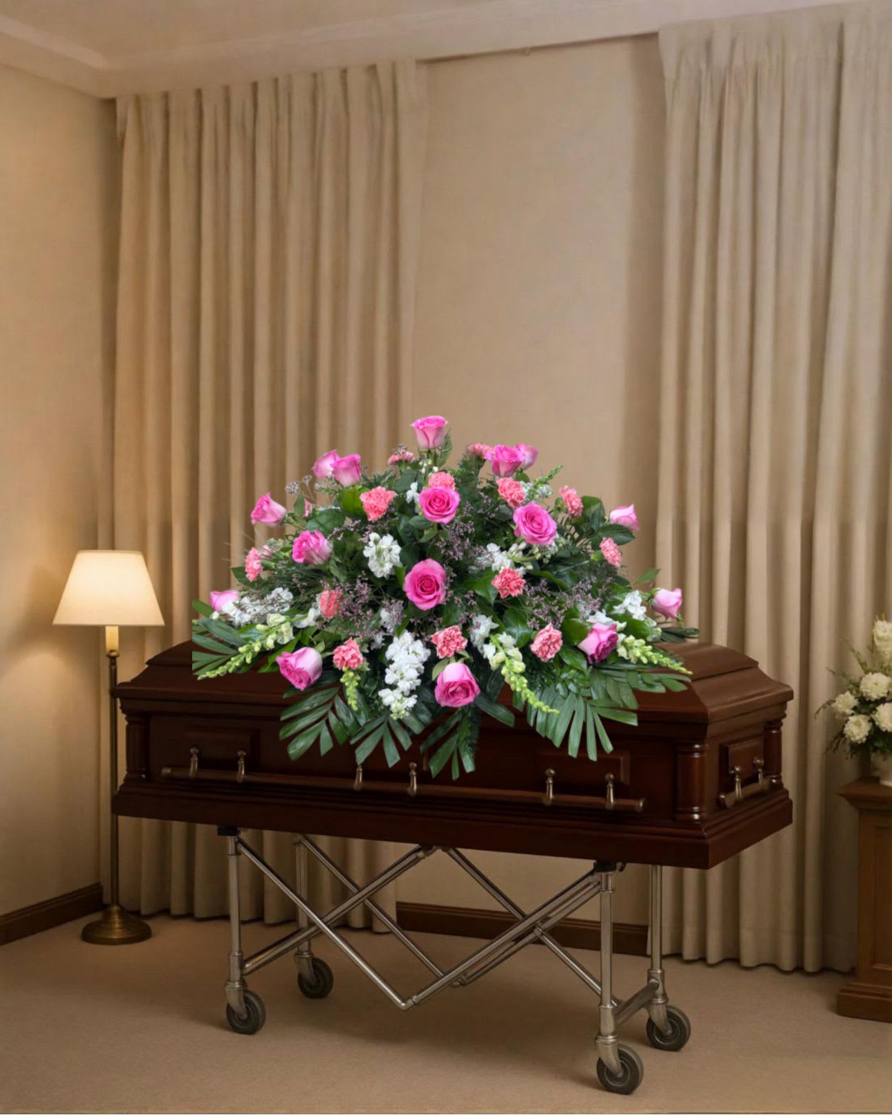 Eternal pink casket spray - A broad, low casket spray that balances vivid pink roses with soft white carnations and delicate pink filler blooms - designed to lay gracefully across a dark wood casket for a funeral or memorial service. The bright pink roses form the focal point so the person you're honoring will be remembered with warmth and dignity, while trailing greenery and palm-like fronds soften the silhouette for a peaceful, respectful presentation.  Crafted to cover the top of the casket, this arrangement features clusters of roses, carnations and mixed filler flowers in pink, white and green tones with lush greenery accents. The spray sits on a metal casket stand and is shown with a floor lamp nearby to indicate scale; proportions are broad and low to create an elegant, reverent display that complements rather than overpowers the service.  Choose this casket spray when you want to convey love and gentle remembrance at the funeral or memorial; it communicates sympathy and celebration of life in equal measure. If you need delivery and placement help, our shop can coordinate casket delivery and set-up to ensure the arrangement is positioned correctly for the service.