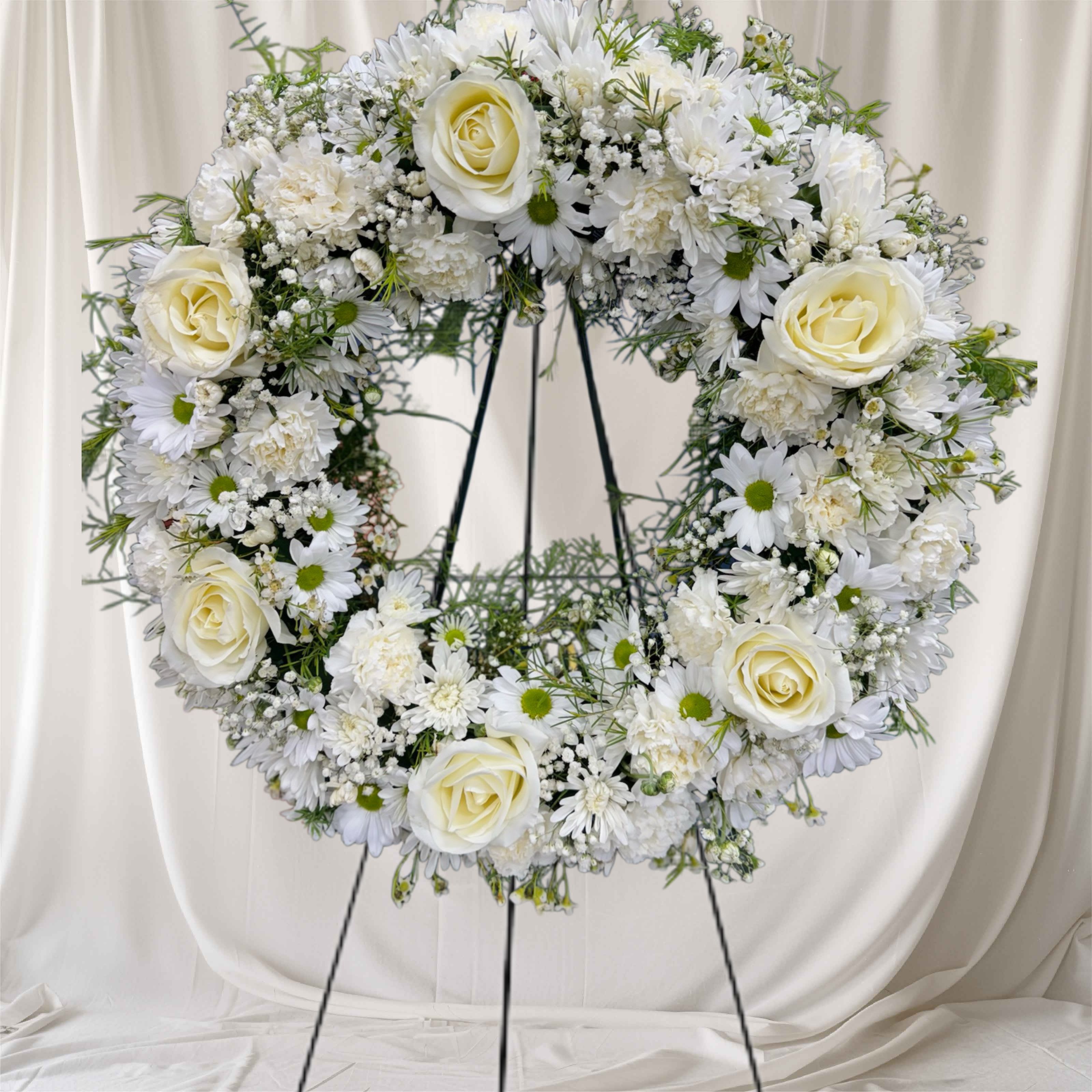 Loving memory  - A soft,18-inch  sympathy wreath that speaks of comfort and remembrance - perfect as a funeral spray or memorial tribute. This loving memory flower arrangement uses a clean palette of white and cream roses, white daisy-like blooms, and white carnations to create a densely rounded heart that reads gentle and reverent; small green centers and fine filler add fresh contrast against the white for a calm, hopeful feel.  Mounted on a sturdy black metal easel stand, the wreath presents beautifully at services or a graveside remembrance, giving the person you've lost a dignified, uplifting display. The classic white tones make this a meaningful way to say you're thinking of them or to honor a life lived; ordering and sizing details are available at checkout to ensure the wreath fits the service space.