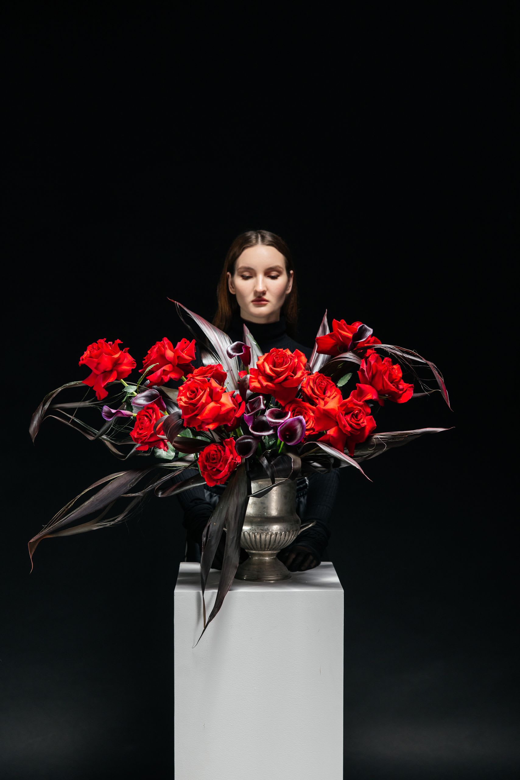 Red Authority Vase Arrangement - Red Authority is not a bouquet—it’s a presence. Velvety red roses command attention at the center, surrounded by sculptural calla lilies and deep, shadow-toned foliage that moves like smoke in the dark. Designed with sharp intention and dramatic balance, this arrangement feels more like a fashion editorial than florals. It’s for bold apologies, unapologetic statements, or moments when “just flowers” isn’t enough. This piece speaks power, control, and desire—without saying a word.
