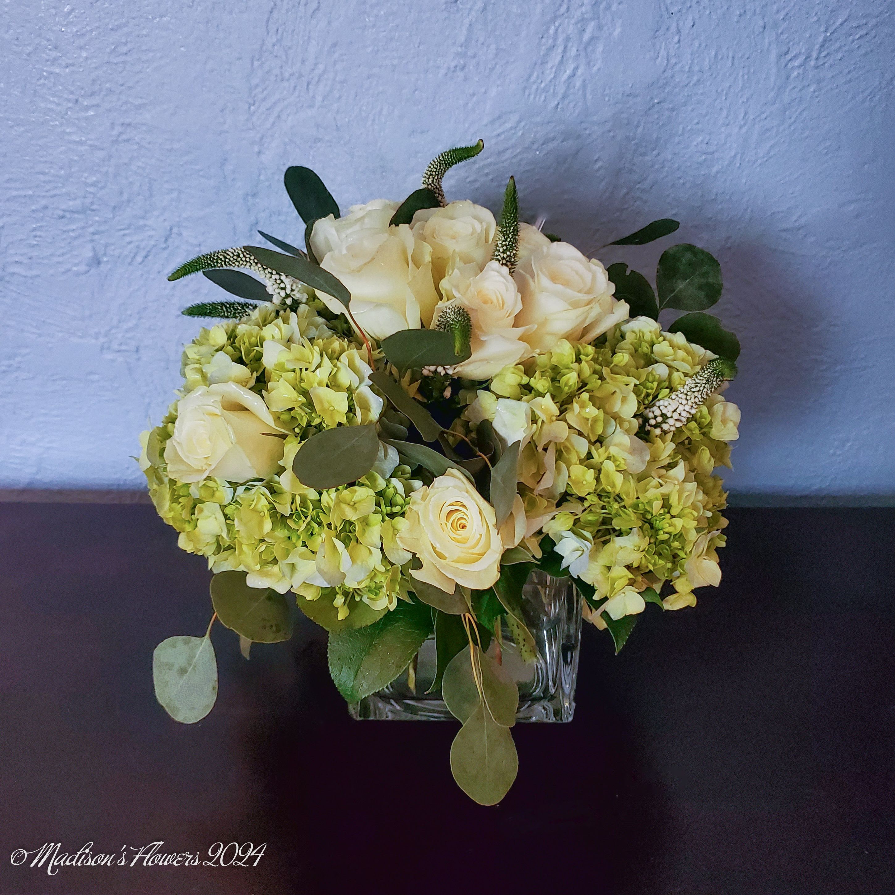 'Simple Classic Greens and Creams' by Madison's Flowers - Classic round table-top arrangement featuring cream colored roses, accent flowers in soft white and green shades, expertly arranged with matching seasonal foliage for any table-top or sideboard. As the roses open, this style steps up the floral drama. Easy to care for and long-lasting. 