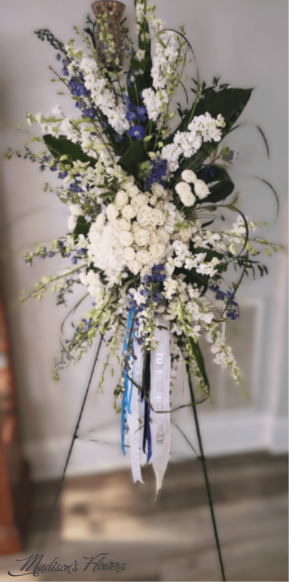 'Remembering Skye, in Bloom' by Madison's Flowers - Our beloved 'Remembering Skye in Bloom' standing spray is designed with a collection of white hydrangeas, white roses, white stock, white snaps, white Delphinium orchids, and a touch of blue delphinium . 