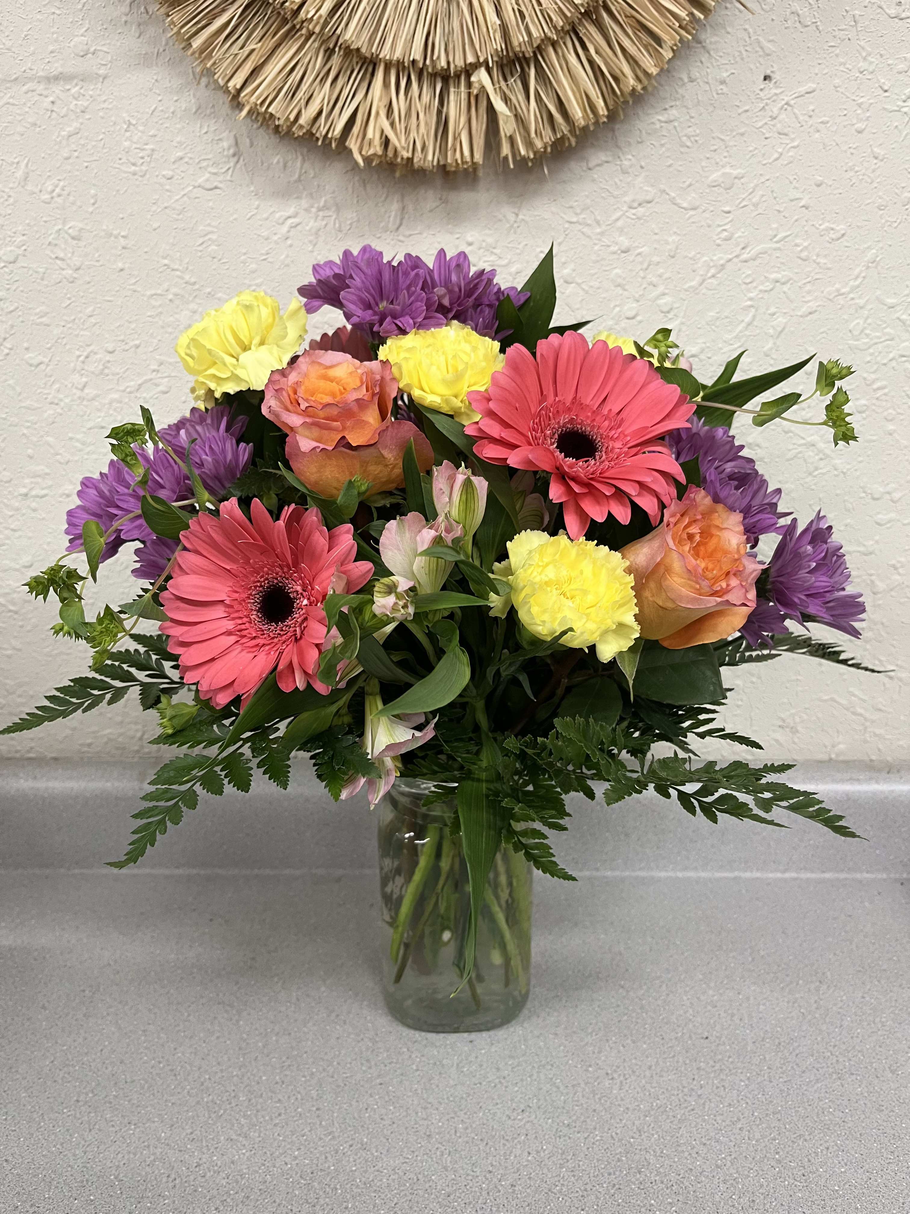 Glamorous Garden - Bright, playful bursts of coral gerbera daisies and free spirit roses make Glamorous Garden perfect for a birthday, Mother's Day, or "just because" surprise. The rounded bouquet reads like a celebration in bloom. They'll feel instantly noticed and uplifted when these vivid pinks and purples meet sunny yellows and warm orange tones, making it a cheerful pick for anniversaries, graduations, or to say "I'm sorry" with sincerity.  Layered densely in a clear glass vase, the arrangement mixes alstroemeria, carnations, gerbera daisies, roses and daisies with rich green foliage for texture and depth. The blooms are arranged at varied heights to create a full, rounded silhouette. 