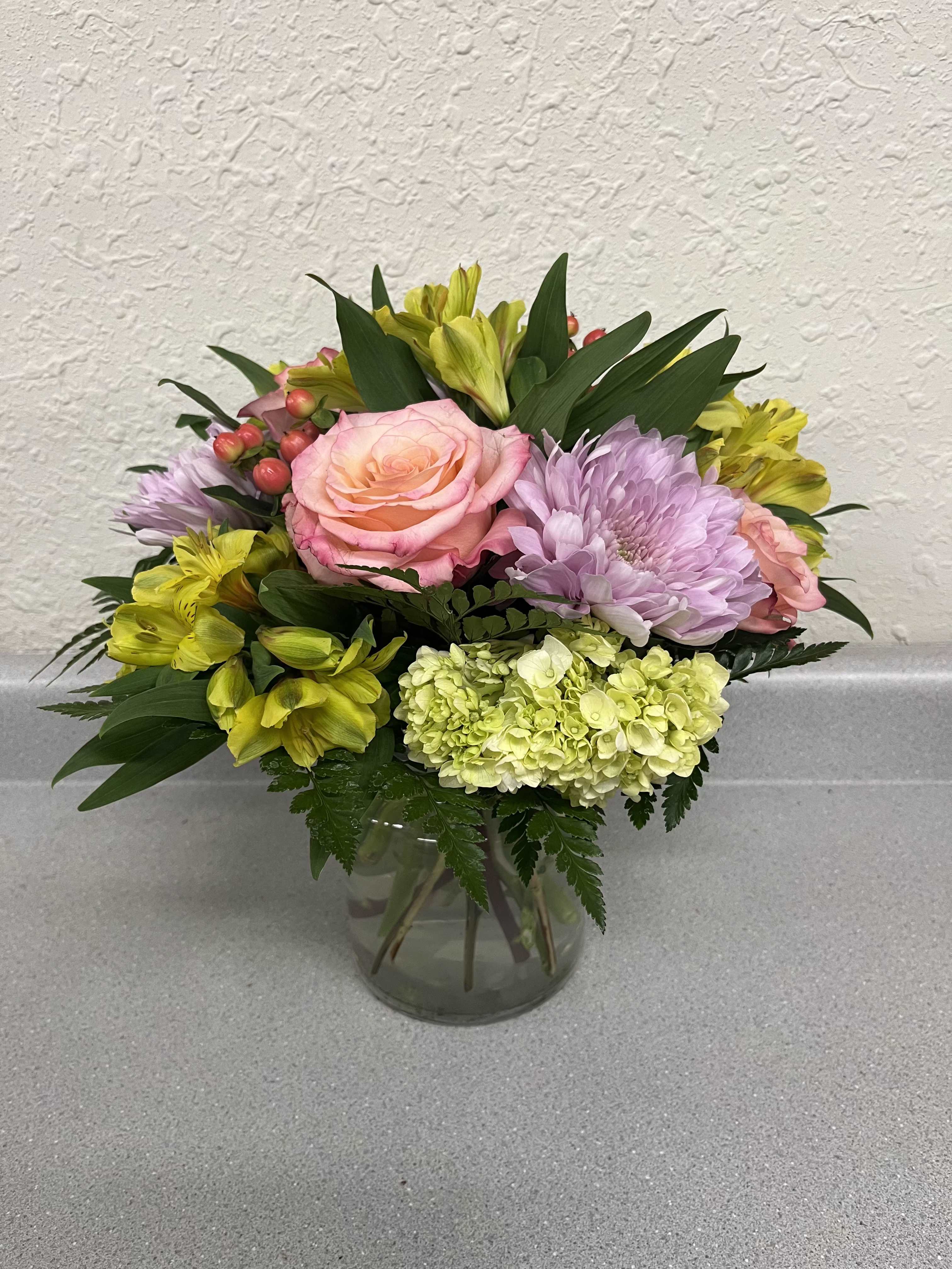 Loving Thoughts - A rounded cluster of pink roses and a pale green hydrangea creates an immediate feeling of warmth and tenderness which is perfect for Mother's Day gift, an anniversary bouquet, or a "thinking of you" surprise. Loving Thoughts is a flower arrangement that is fresh and cheerful with soft pinks and lavenders meeting bright yellow alstroemeria for a lift.  This arrangement combines roses, lavender chrysanthemums, yellow alstroemeria, and a green hydrangea tucked into a clear glass vase, layered densely with broad dark foliage for contrast and fullness. Small coral berry accents add playful texture and visual interest. Sized to sit nicely on a table or desk, it suits housewarmings, retirement celebrations, get-well wishes, and everyday expressions of love and appreciation.