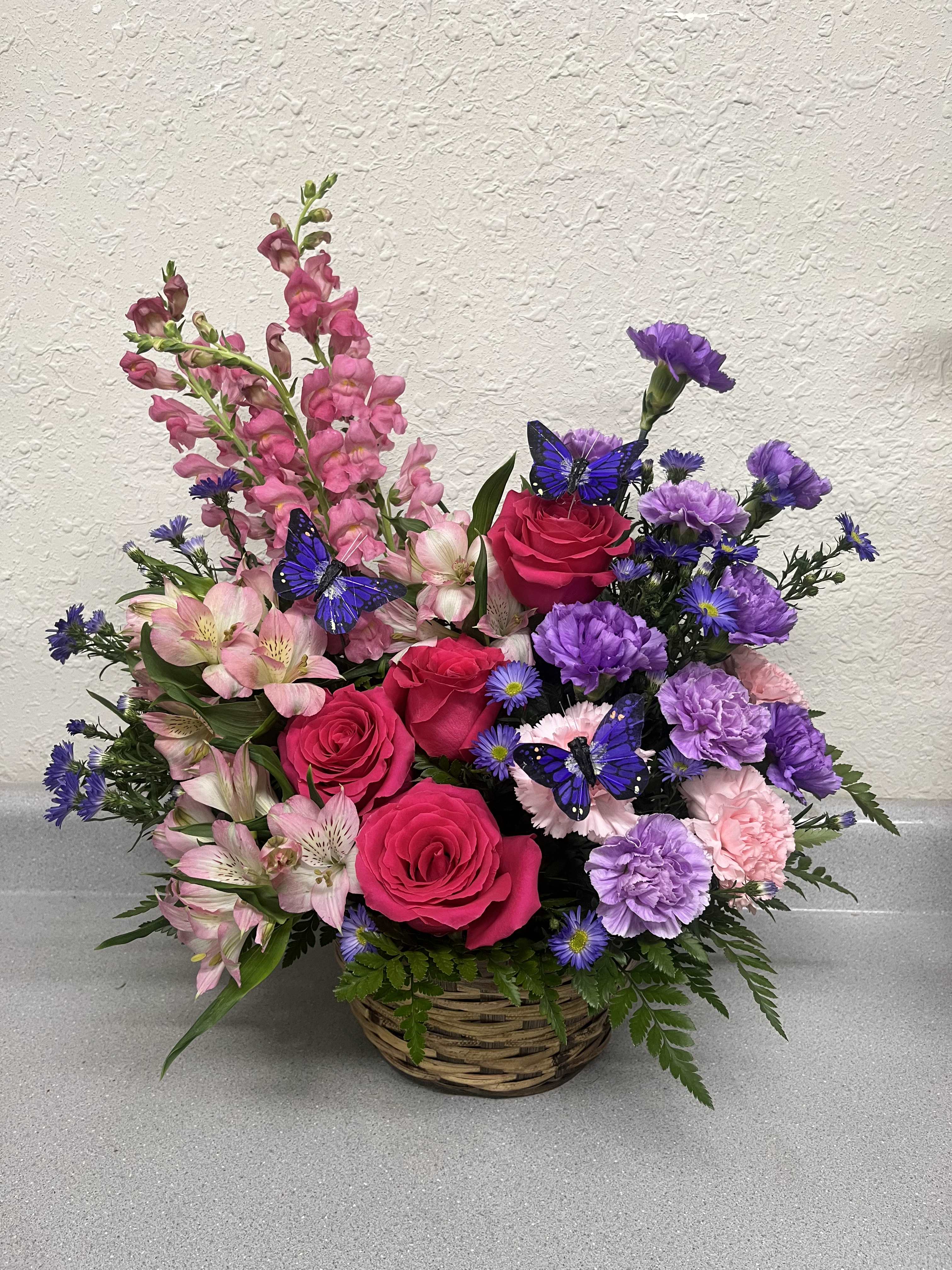 Basket arrangement of pink roses, purple carnations, and pink snapdragons with butterfly picks